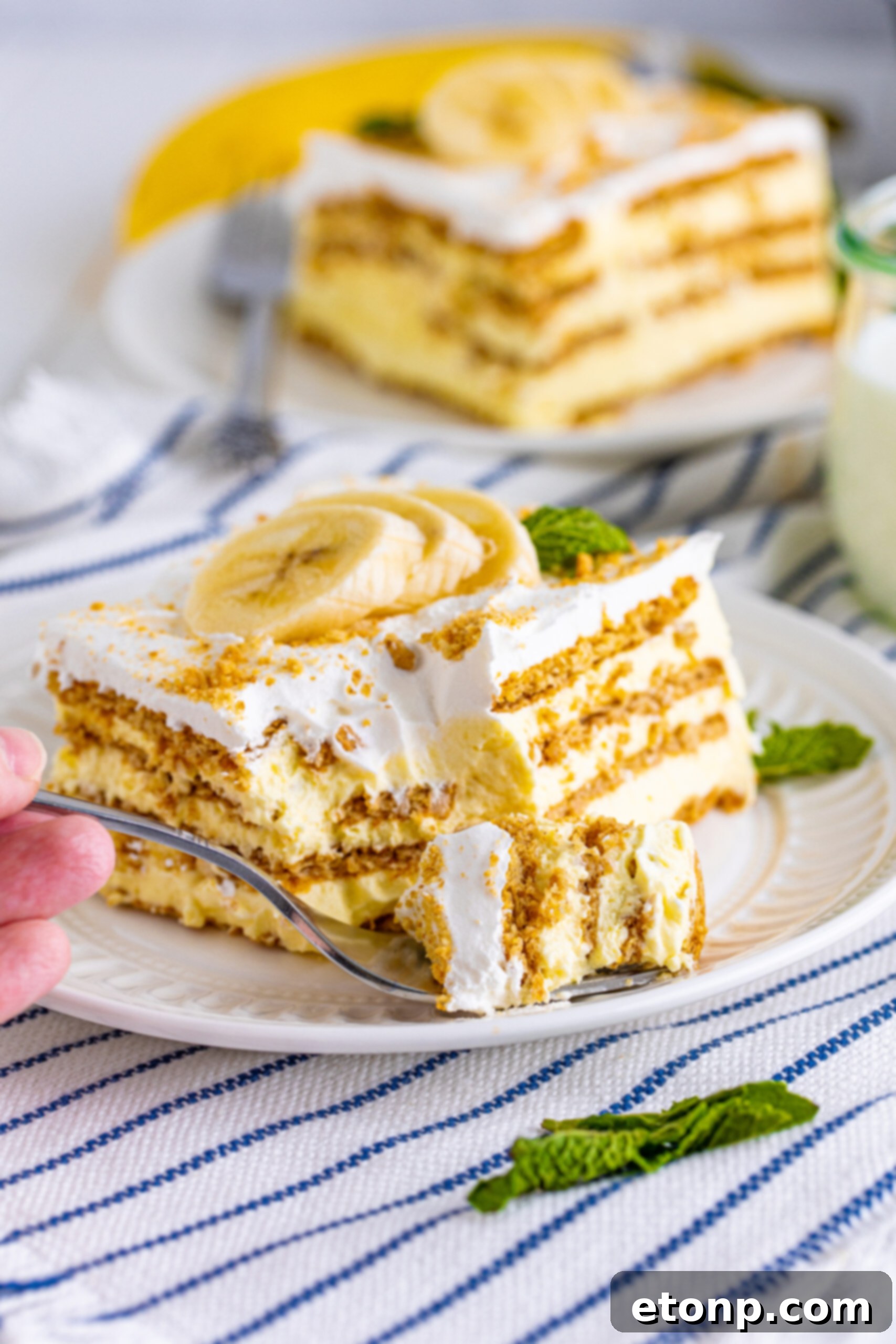 A slice of Banana Icebox Cake on a white plate, with a silver fork having taken a bite, highlighting the moist and creamy texture of the dessert.