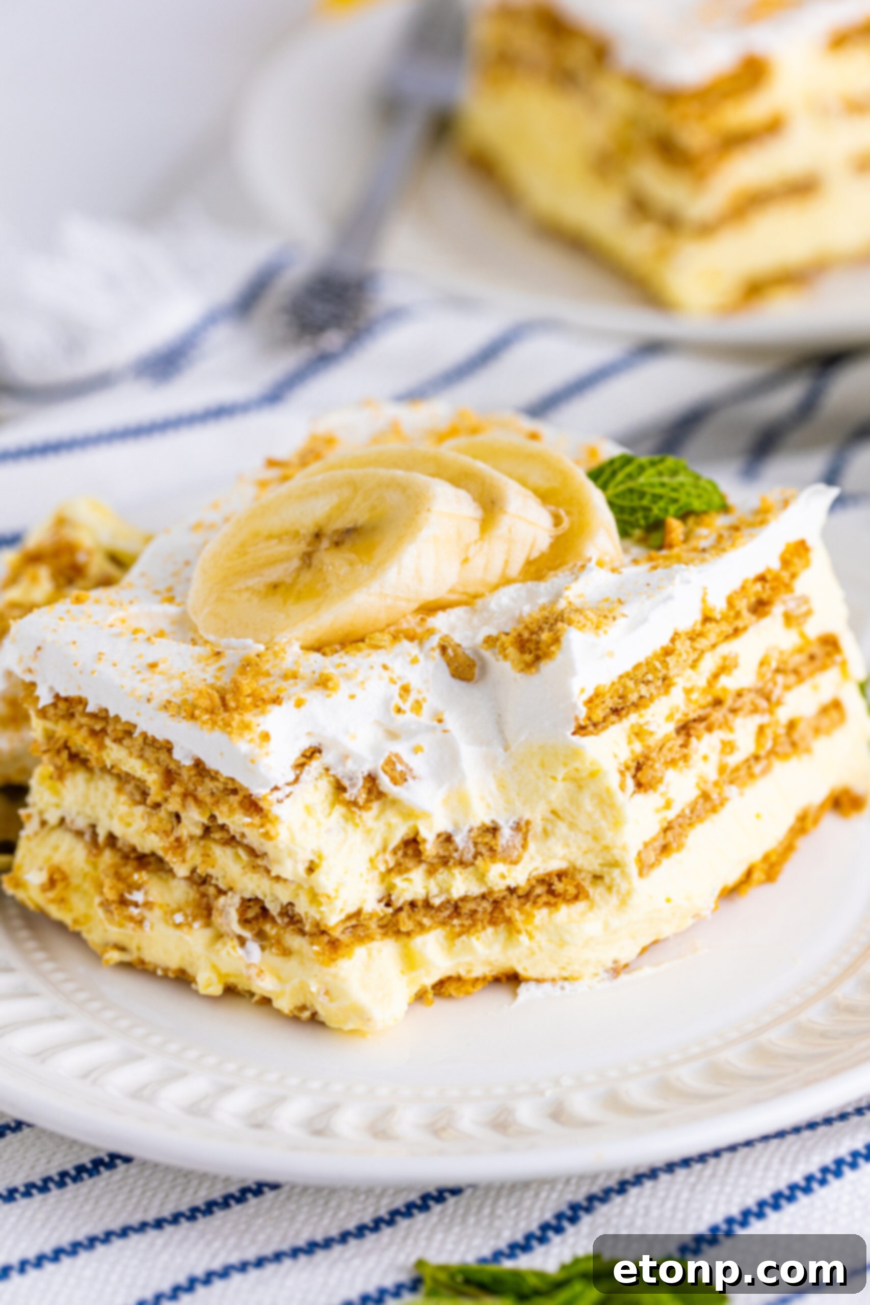 Another perspective of a Banana Icebox Cake slice on a white plate, showing a bite removed by a silver fork, emphasizing its tender texture.