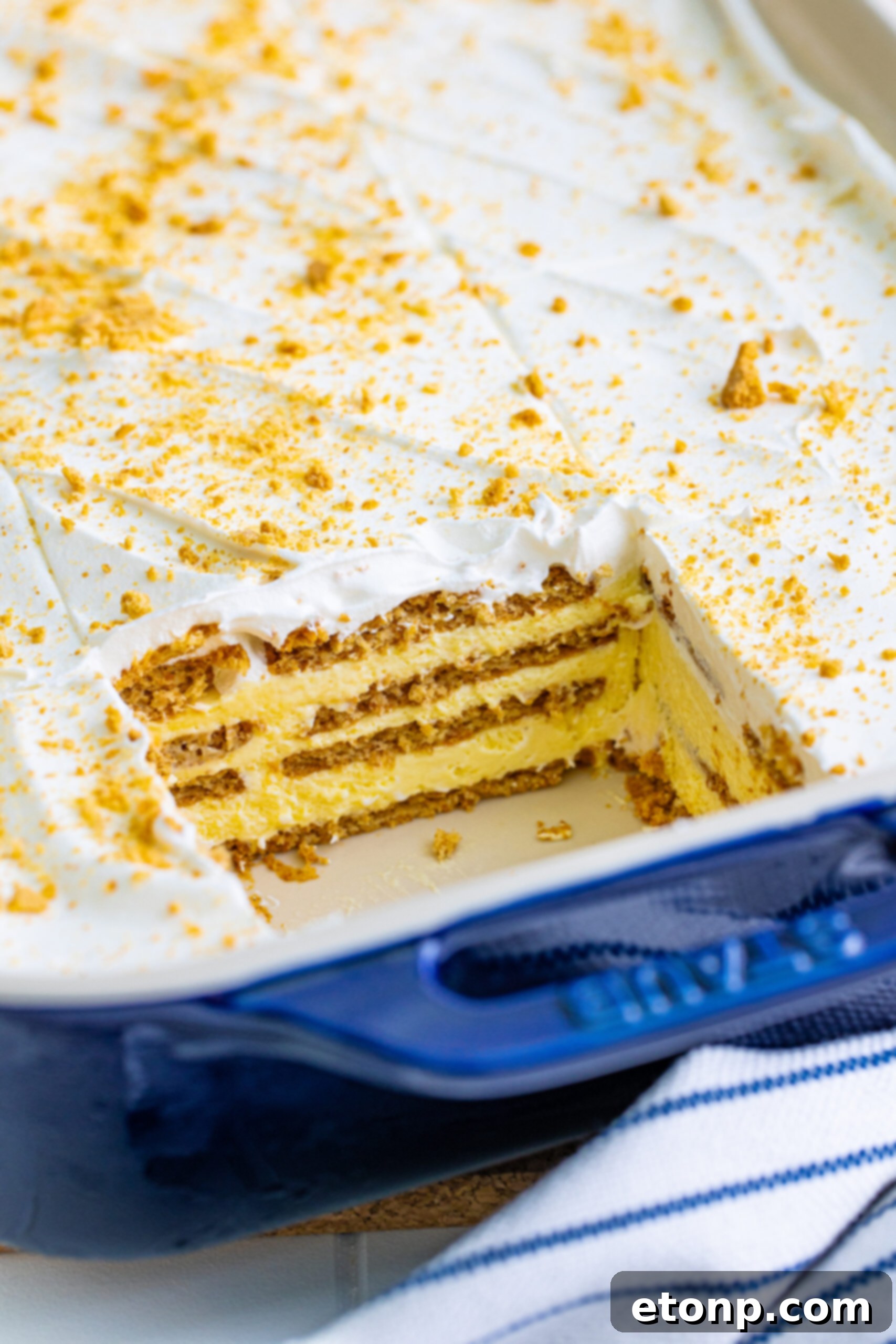 A vibrant blue baking dish holds a complete No-Bake Banana Icebox Cake, with one slice already removed, revealing its inviting interior. The top is generously garnished with crushed vanilla wafers.