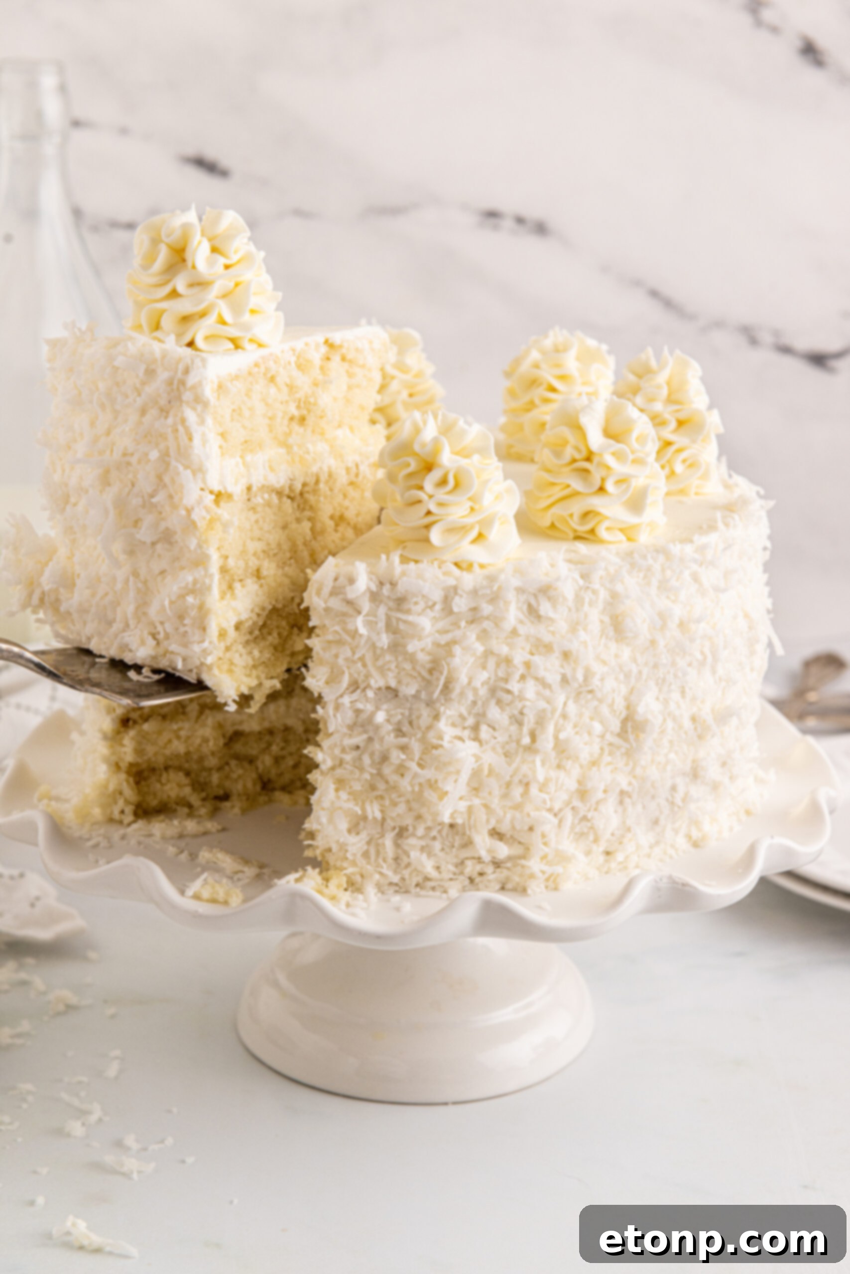 Front view of Coconut Layer Cake on a white cake stand with a slice being removed by a silver cake server, revealing its moist layers and creamy frosting.