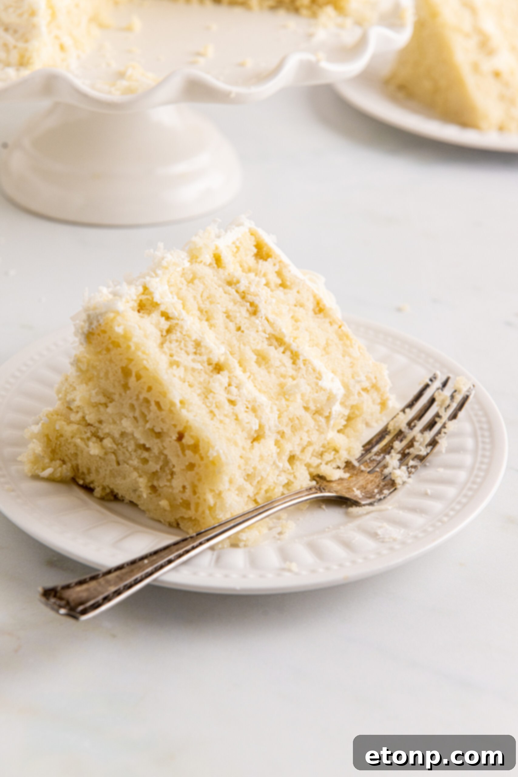 Front view of a slice of Coconut Layer Cake on a white plate with a bite removed, inviting you to taste its perfection.