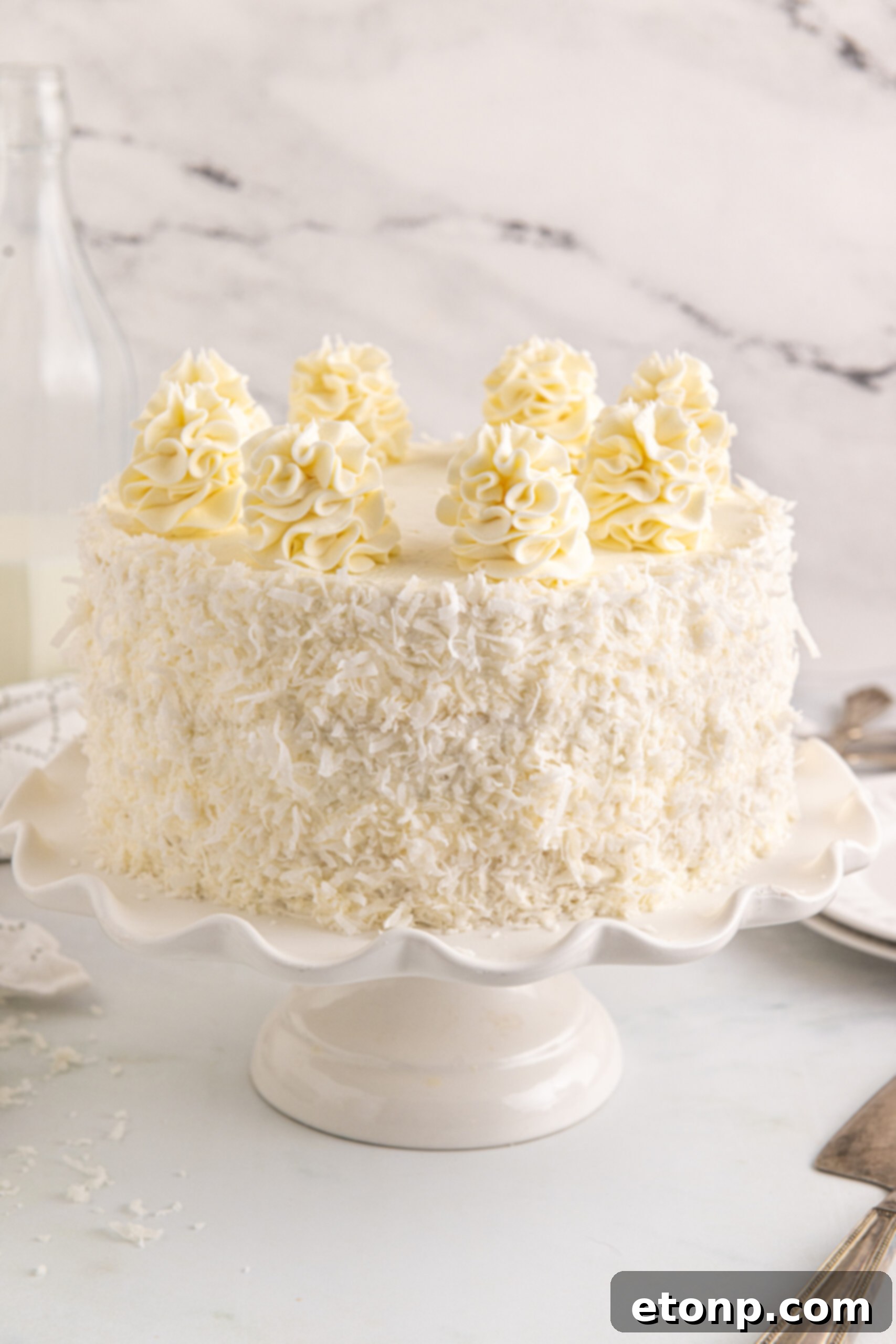 Front view of an elegantly frosted Coconut Layer Cake on a pristine white cake stand, ready to be served.