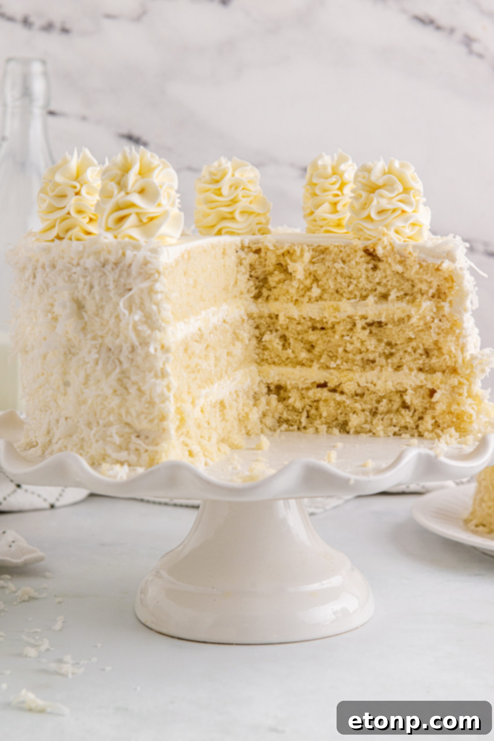 Front view of a beautifully assembled Coconut Layer Cake on a white cake stand with two slices removed, showcasing its delicious layers and frosting.