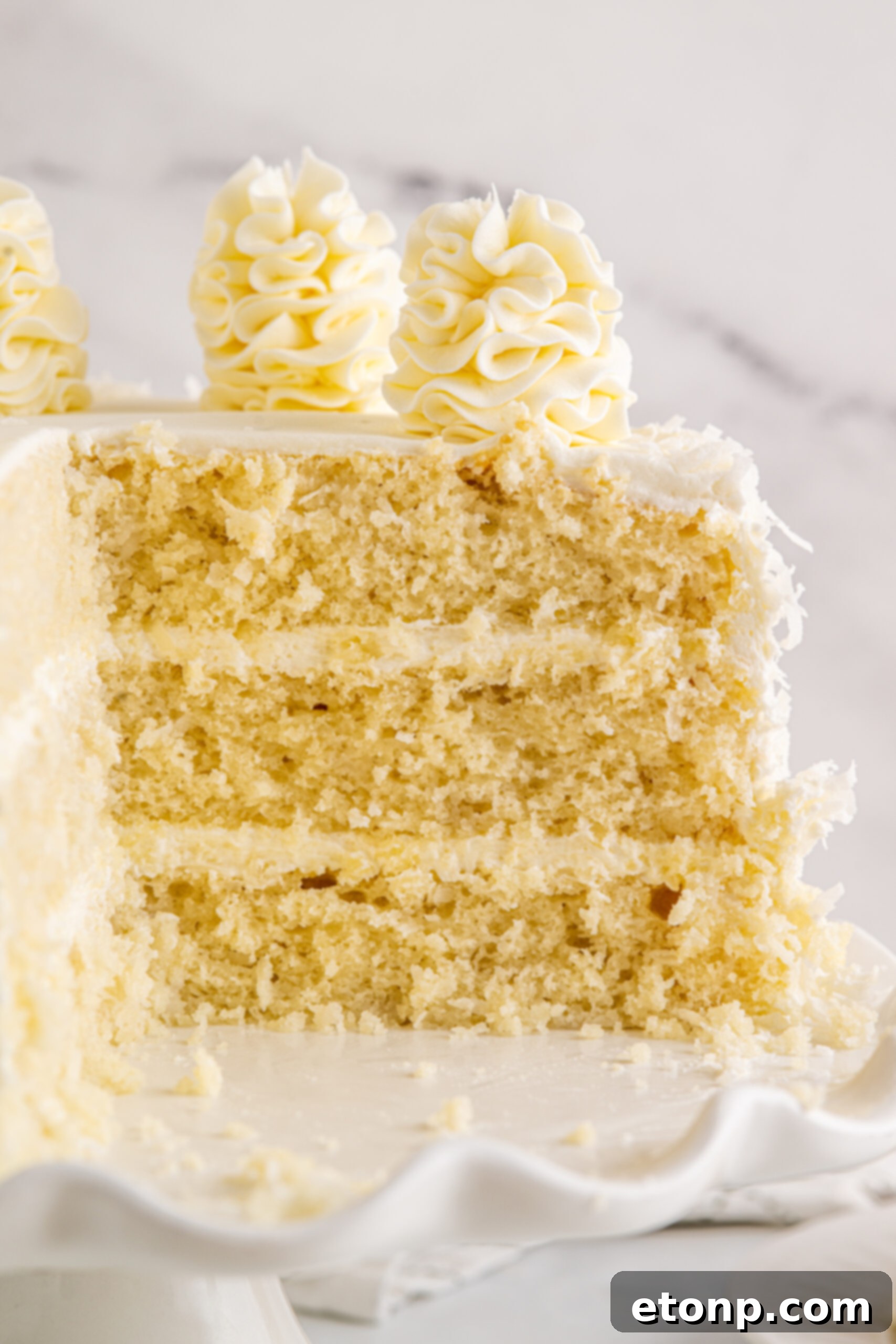 Close up view of the Coconut Layer Cake on a white cake stand with two slices already removed, emphasizing its moist interior and generous frosting.