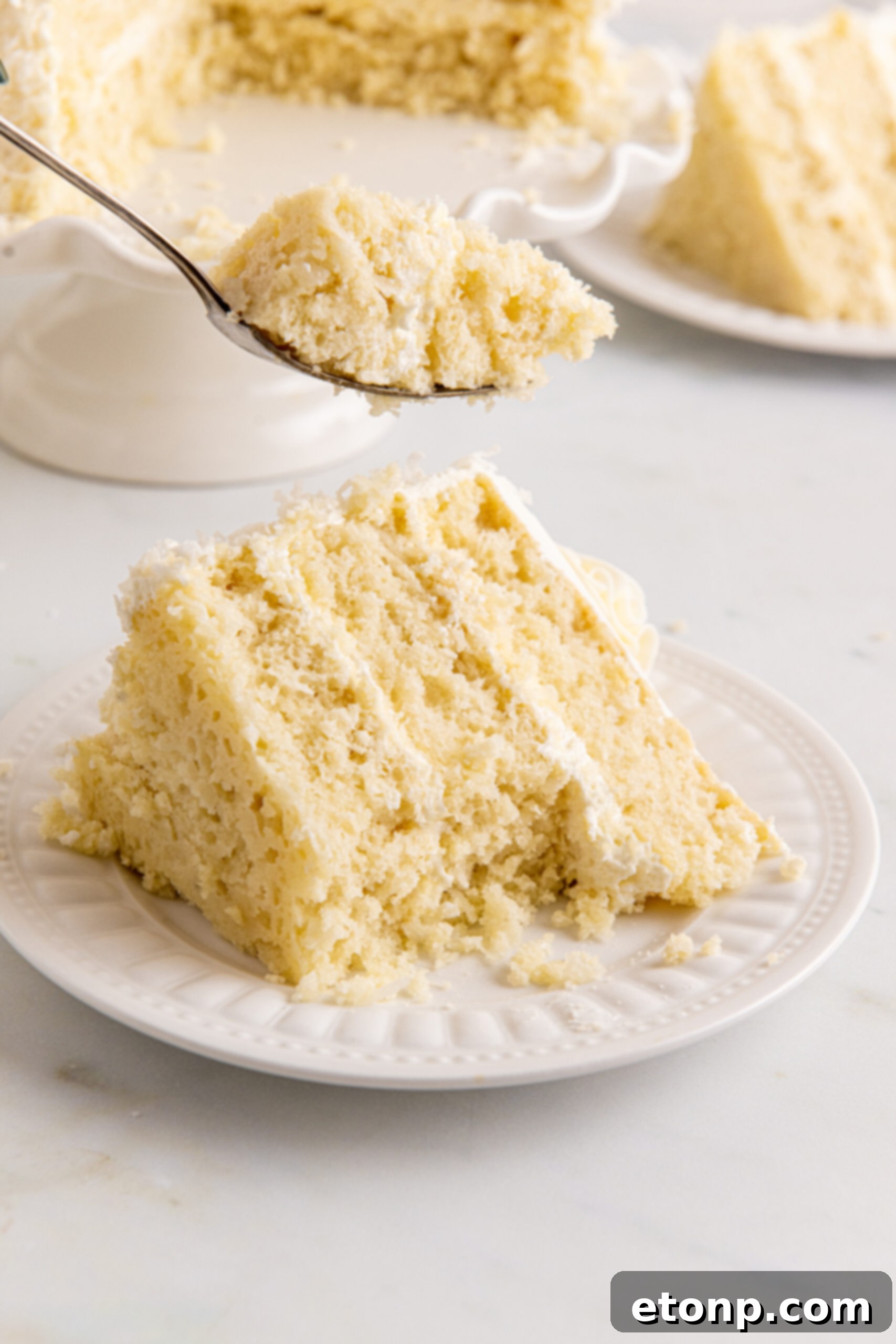 Front view of a slice of Coconut Layer Cake on a white plate with a bite removed by a silver fork, showcasing its delightful layers and tempting texture.