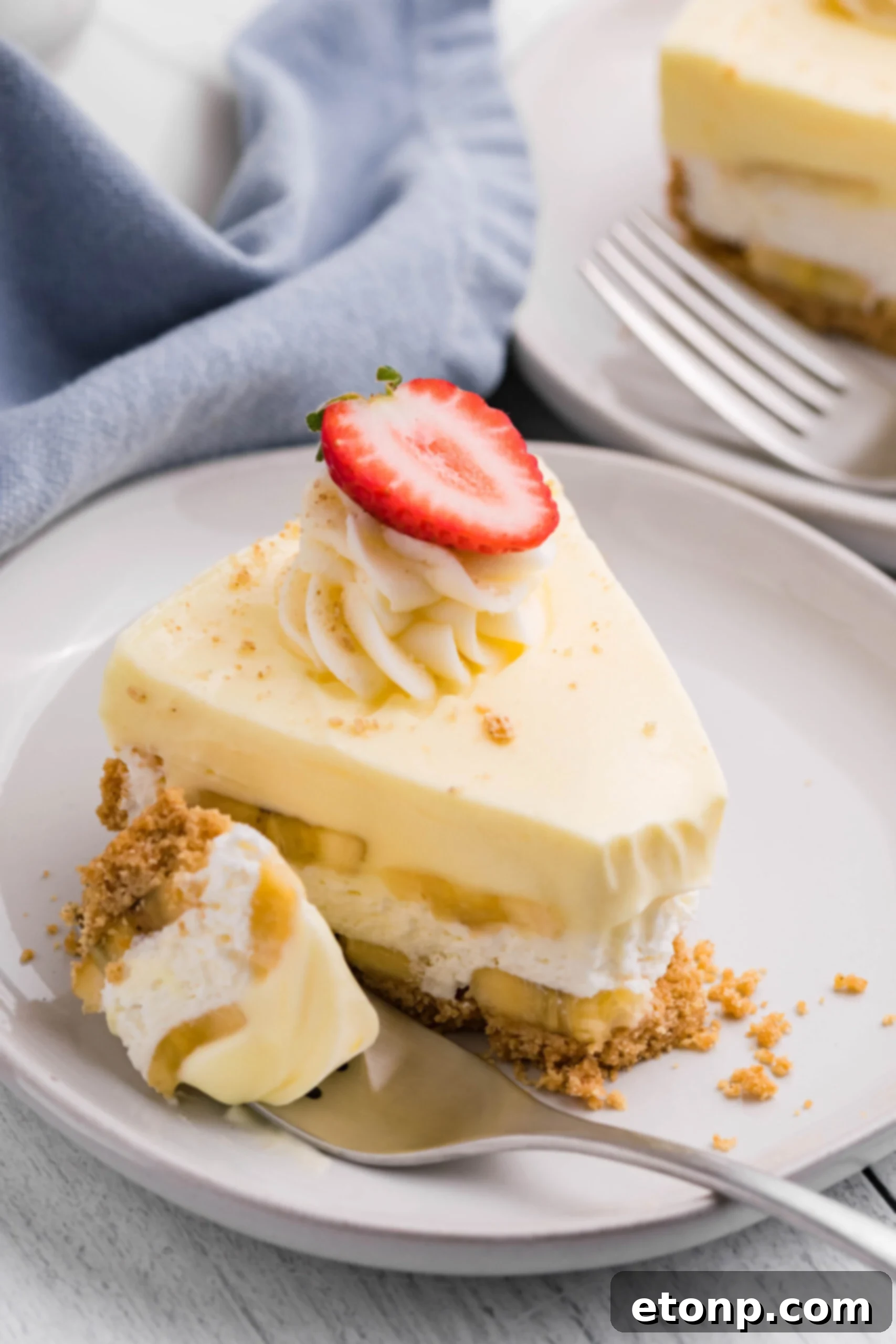 Side view of a slice of Banana Cream Cheesecake on a white plate with a bite removed by a silver fork. A truly decadent and easy summer dessert.