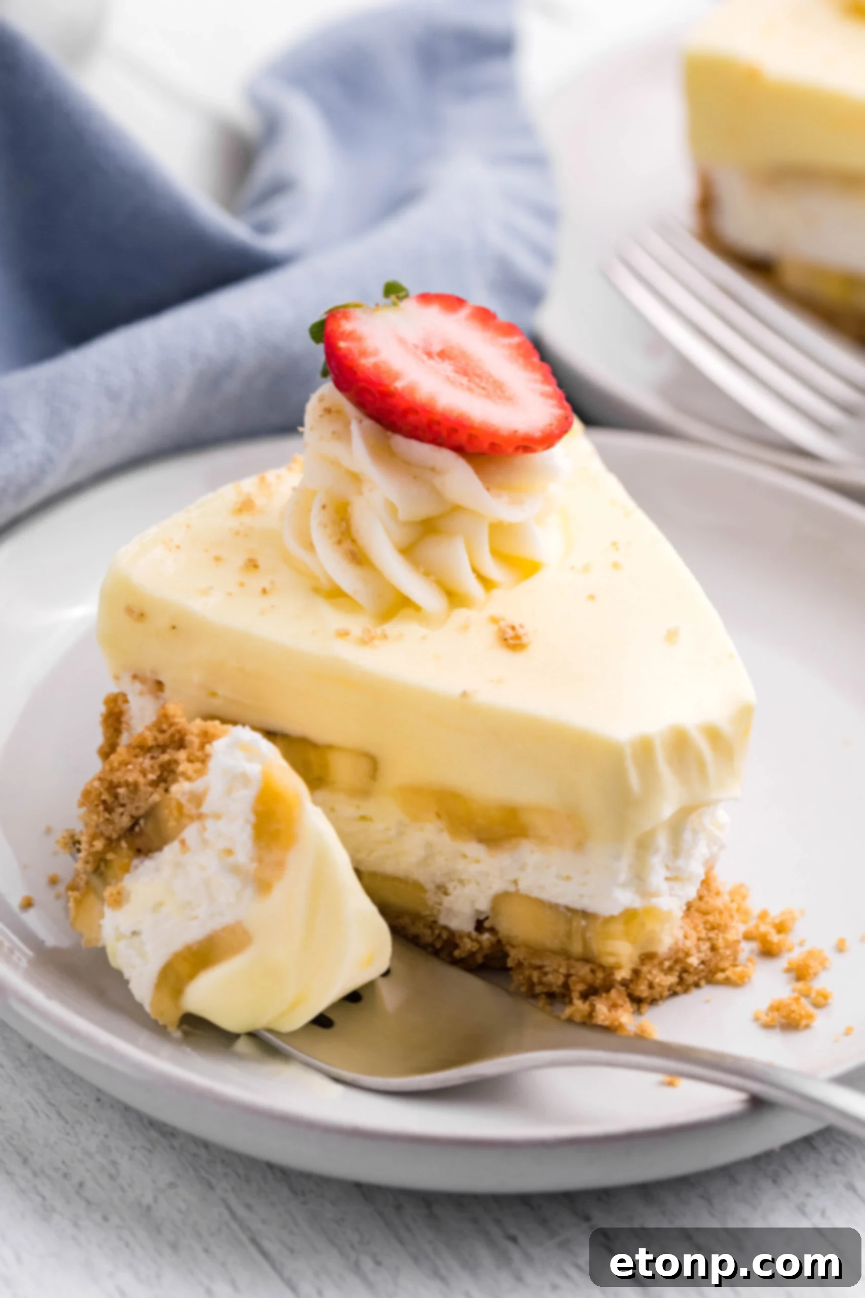 Side view of a slice of Banana Cream Cheesecake on a white plate with a bite removed by a silver fork, highlighting the creamy texture.