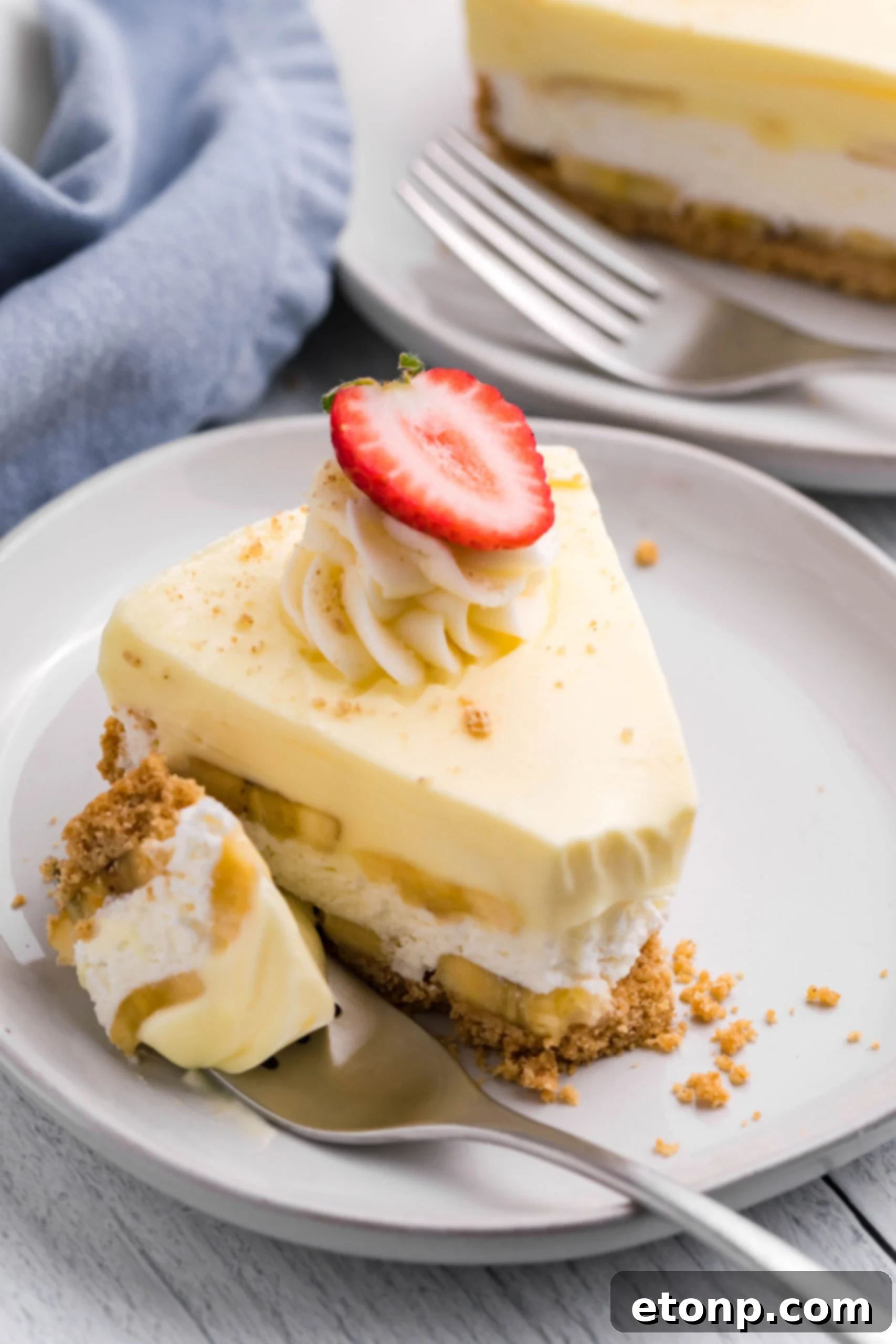 Front view of a slice of Banana Cream Cheesecake on a white plate with a bite removed by a silver fork, showcasing the multiple layers.