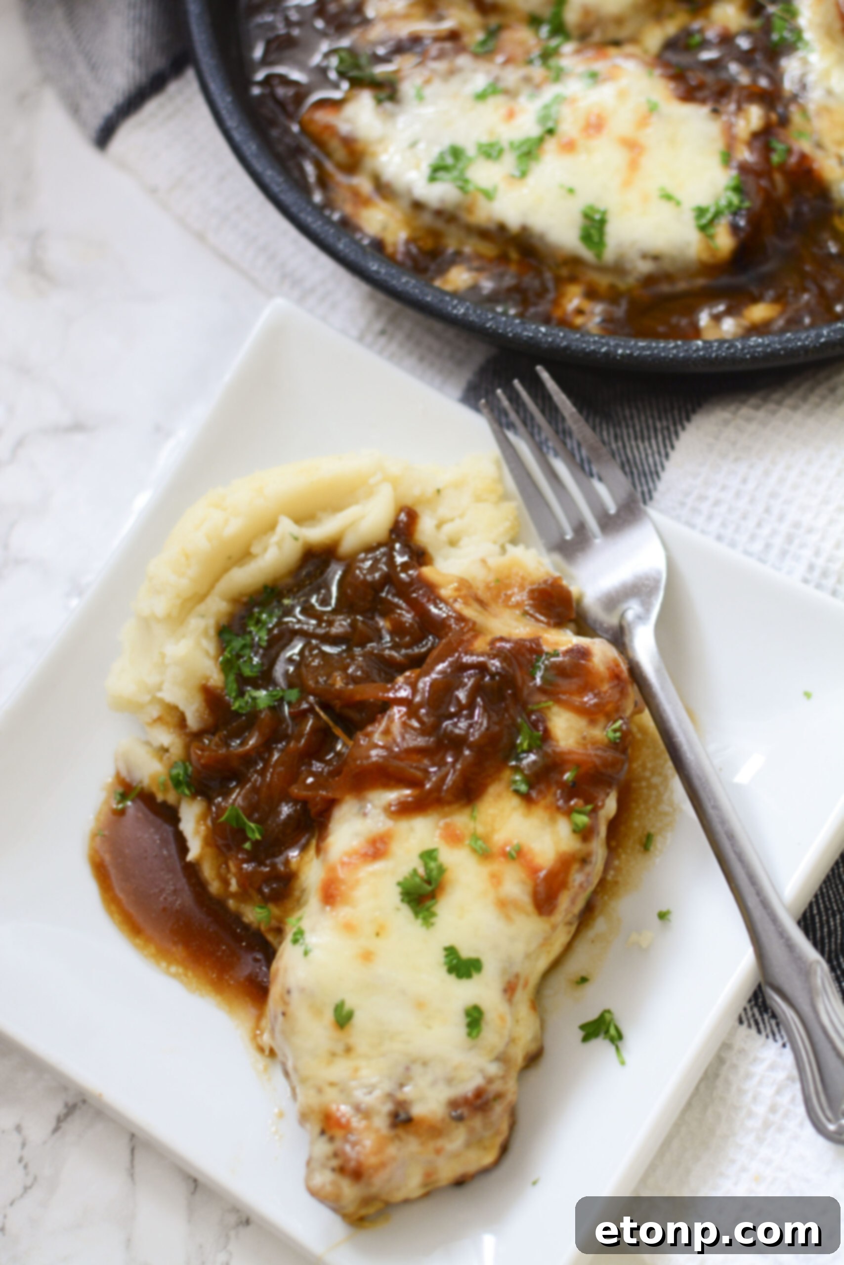 Overhead view of a white plate holding a French Onion Pork Chop, generously coated in gravy, accompanied by creamy mashed potatoes and a silver fork.