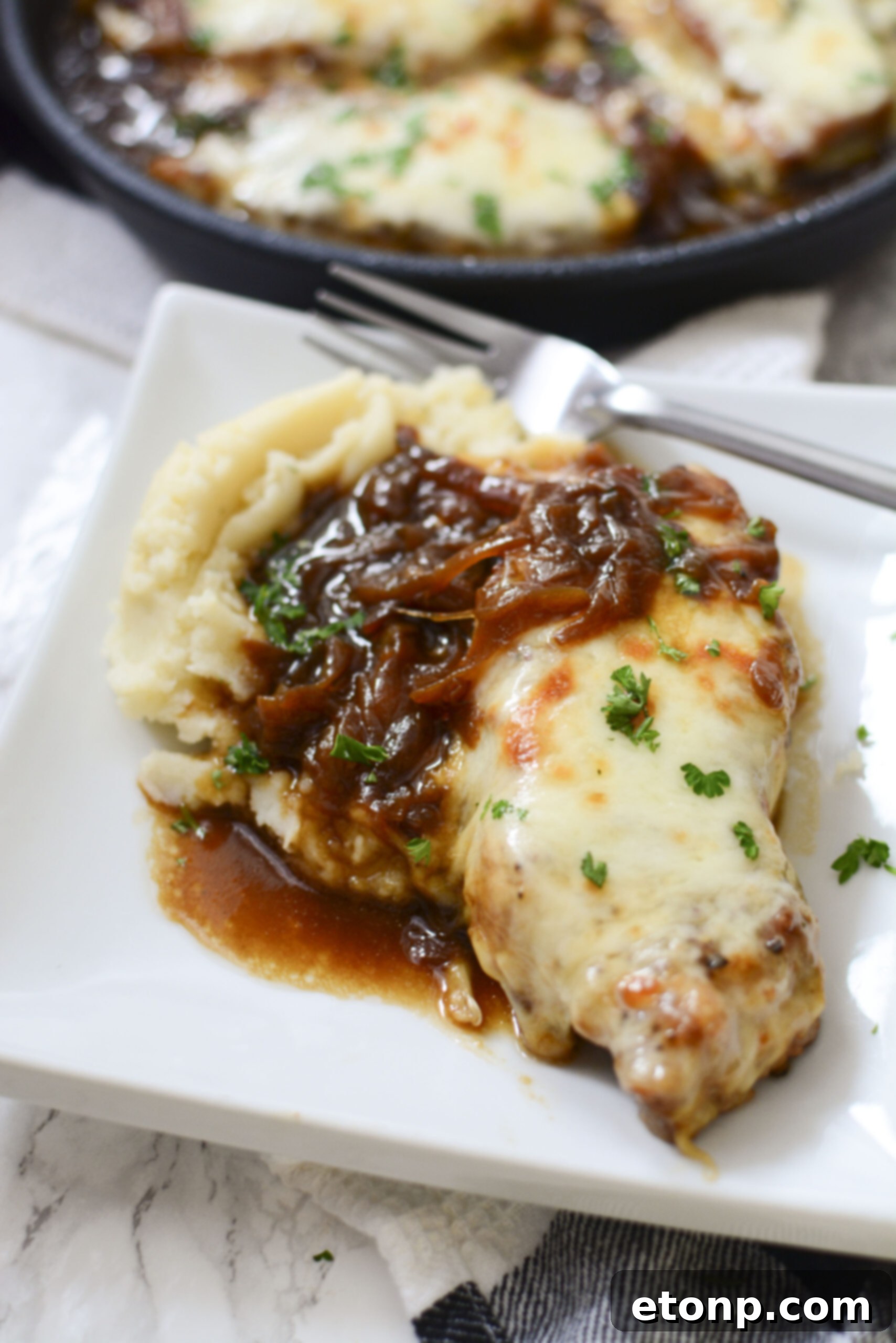 Close-up view of a French Onion Pork Chop on a white plate, served with mashed potatoes, showcasing its rich gravy and perfectly melted cheese.