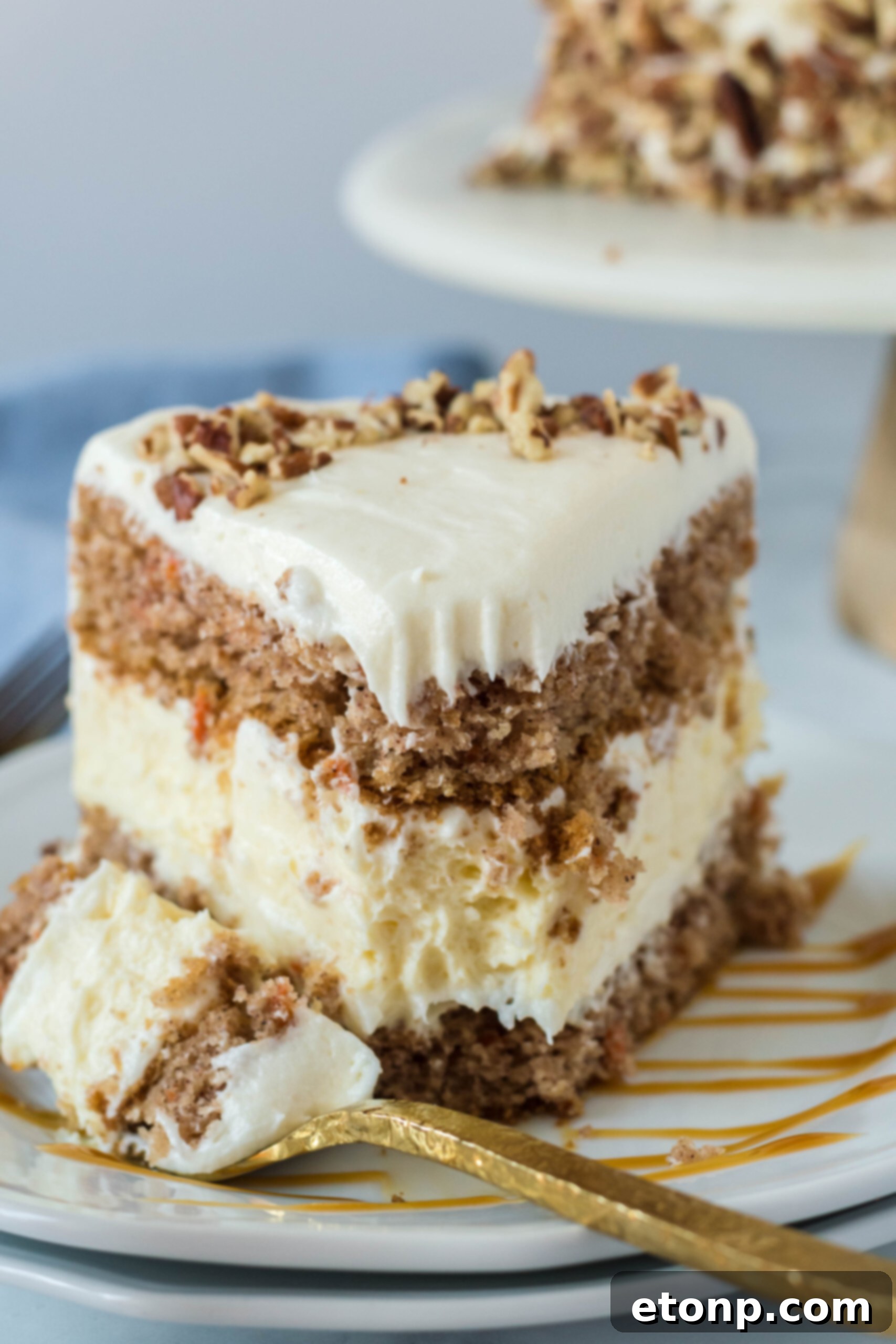 Front view of a slice of Carrot Cake Cheesecake with a bite removed by a gold fork on a white plate drizzled with caramel.