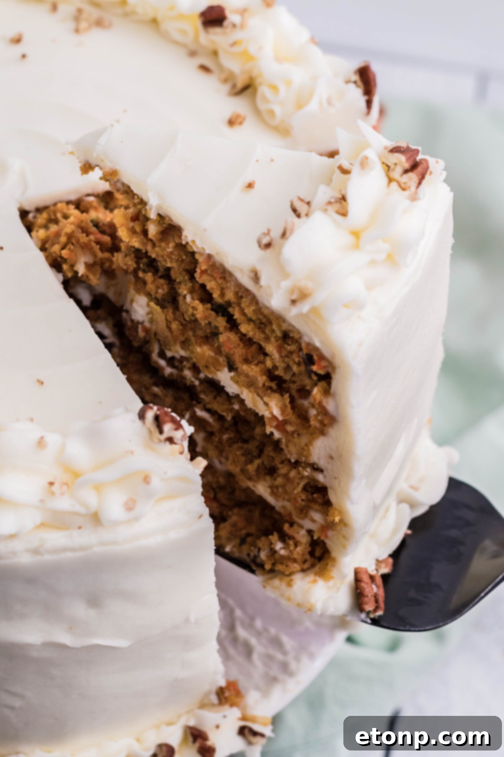 A close-up view of a hand using a black cake server to lift a perfect slice of carrot cake from the main cake, showcasing its layers and rich frosting.