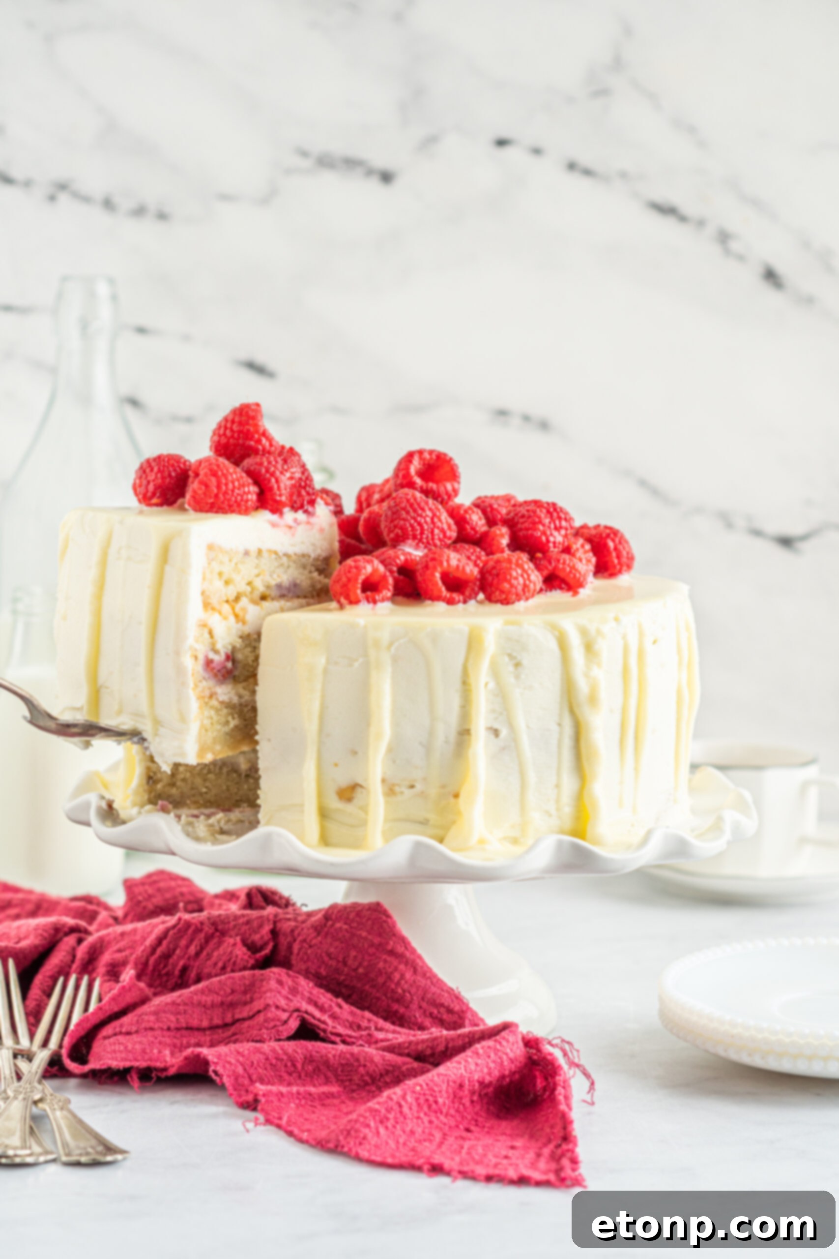 Front view of White Chocolate Cake with Raspberries with a slice being removed by a silver cake server.