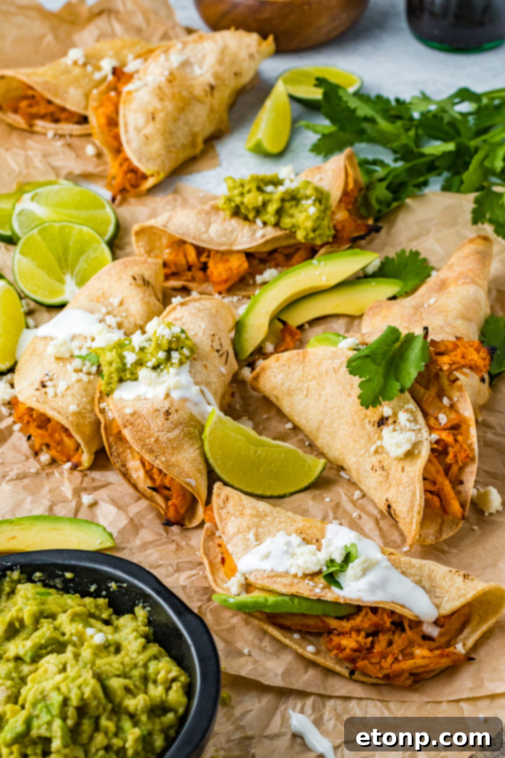 A front view of golden-brown Crispy Tinga Tacos on parchment paper, garnished with fresh lime wedges and sliced avocado, highlighting their inviting texture and presentation.