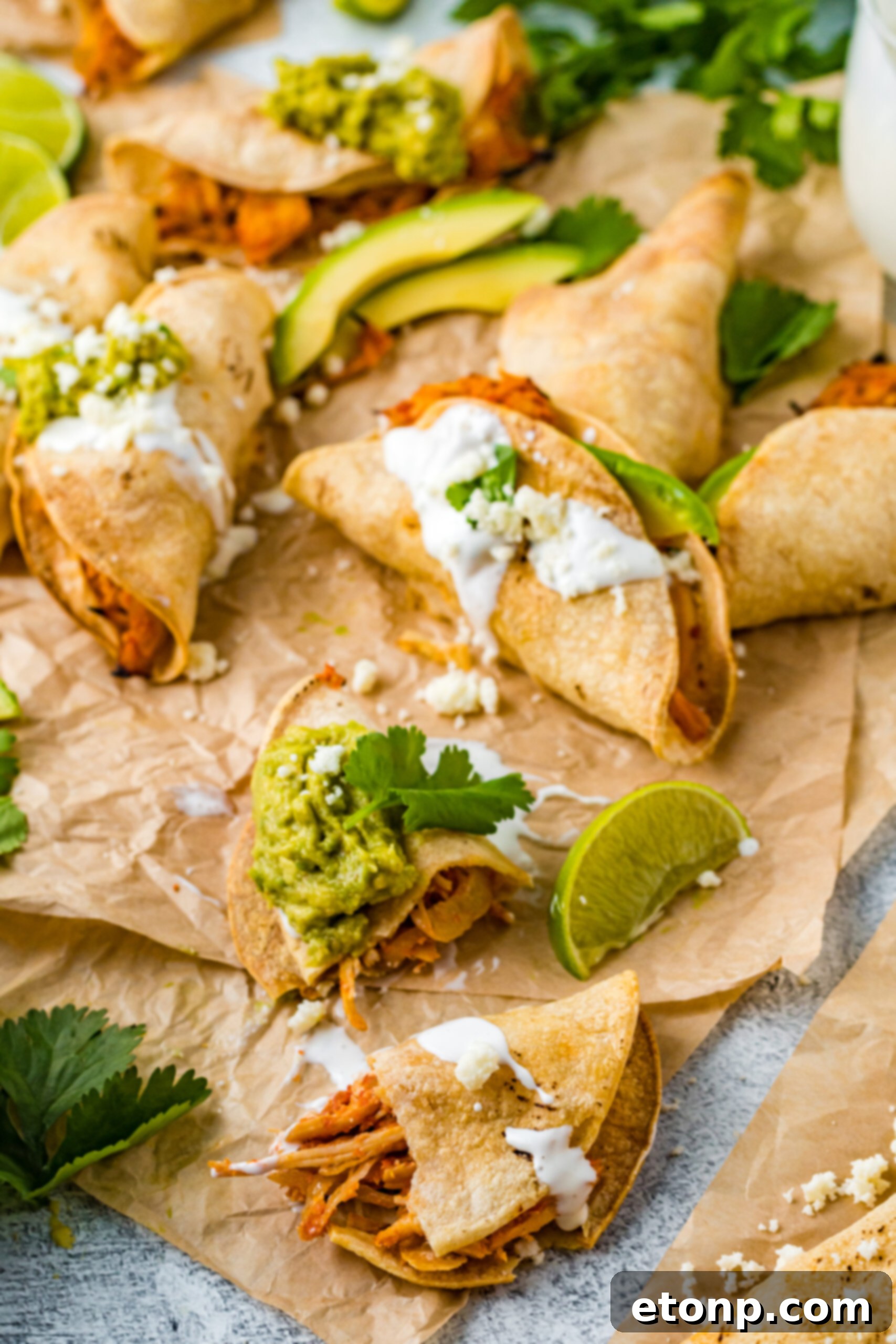 Front view of Crispy Tinga Tacos on parchment paper with lime and avocado slices, featuring one taco split open to reveal its succulent chicken filling.