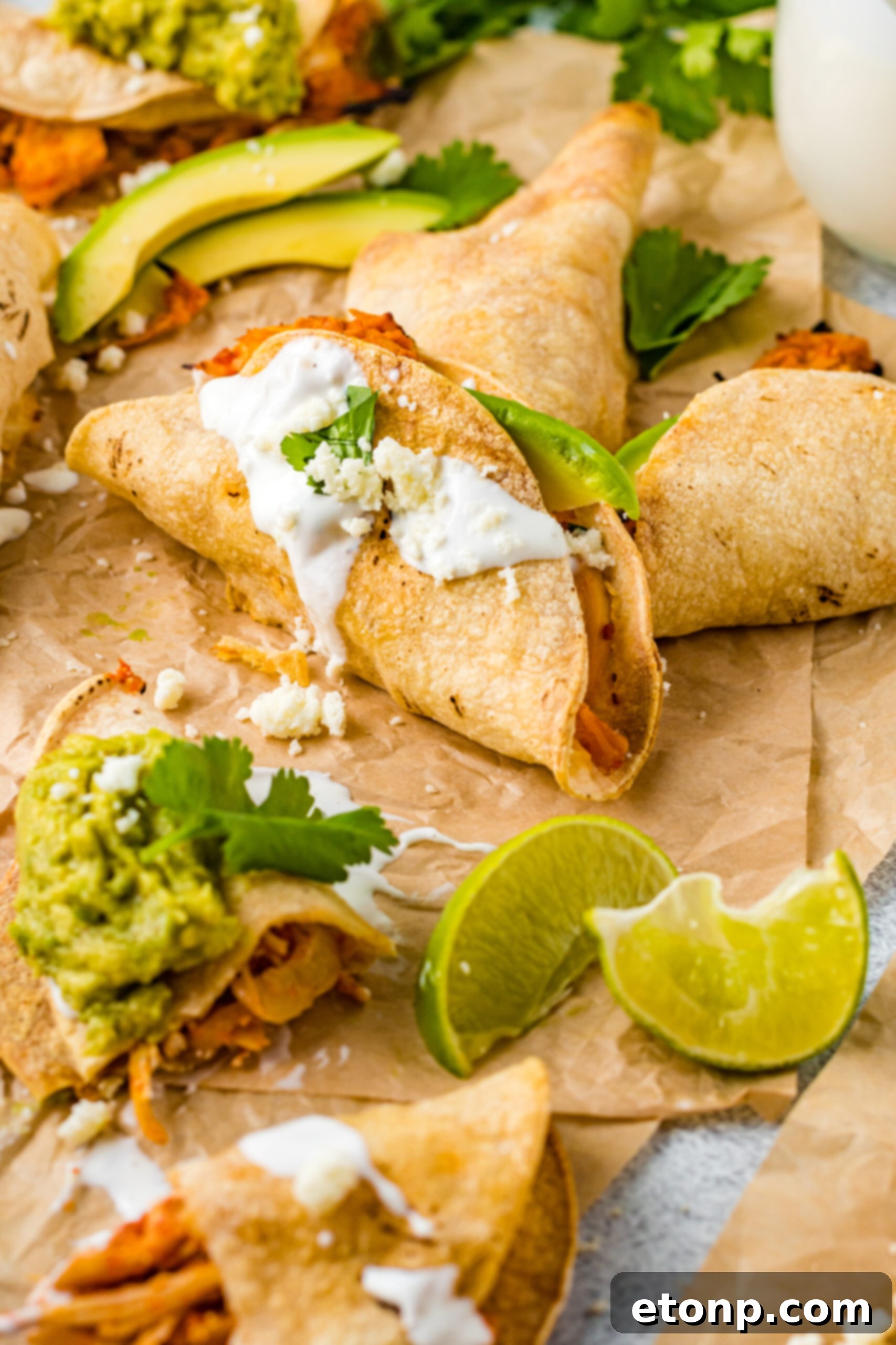 A close-up, appetizing front view of Crispy Tinga Tacos on parchment, with a backdrop of lime and avocado, highlighting their golden crispness.