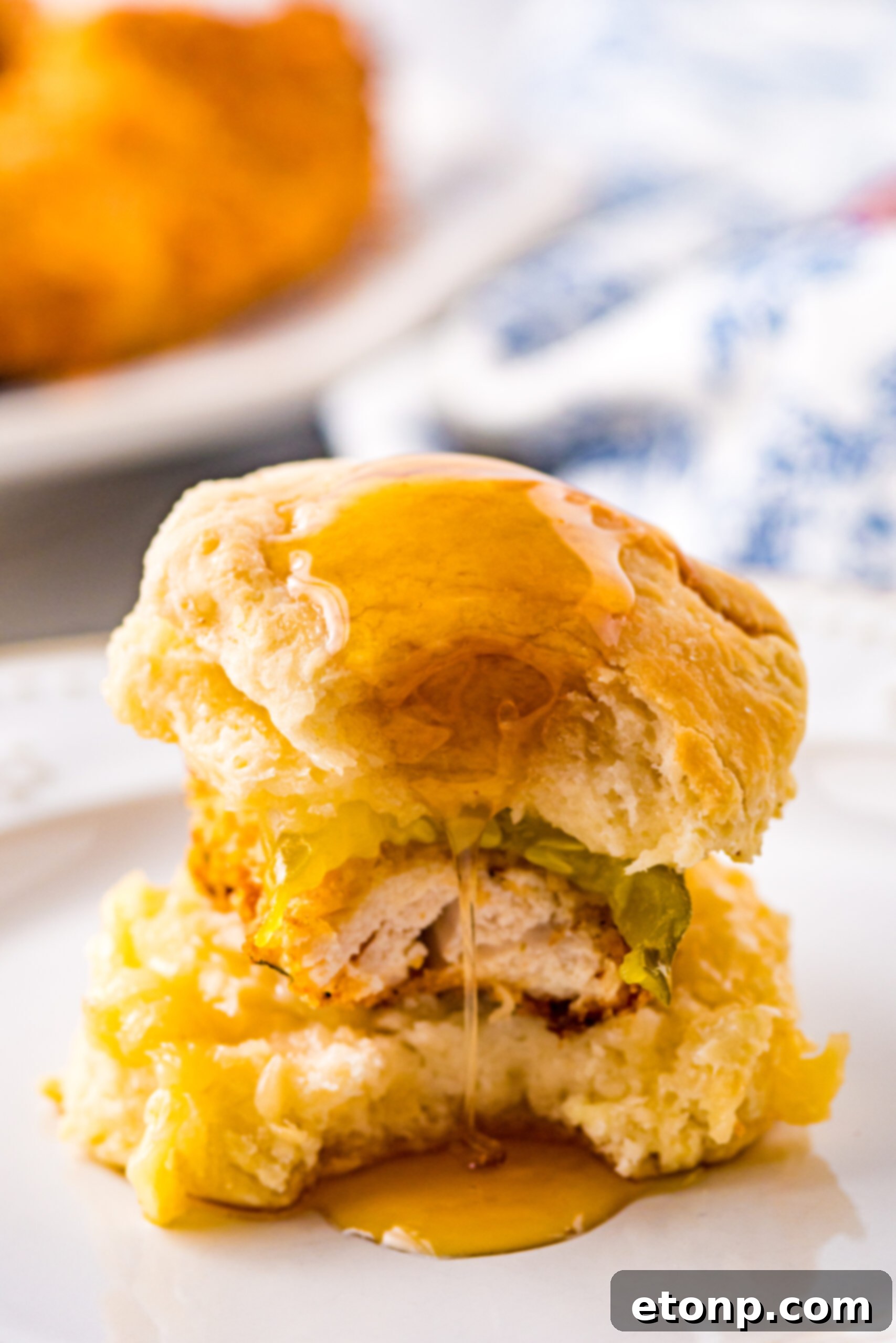 Front view of a Honey Butter Chicken Biscuit on a white plate with a bite removed, revealing the delicious layers inside and inviting you to take a bite.