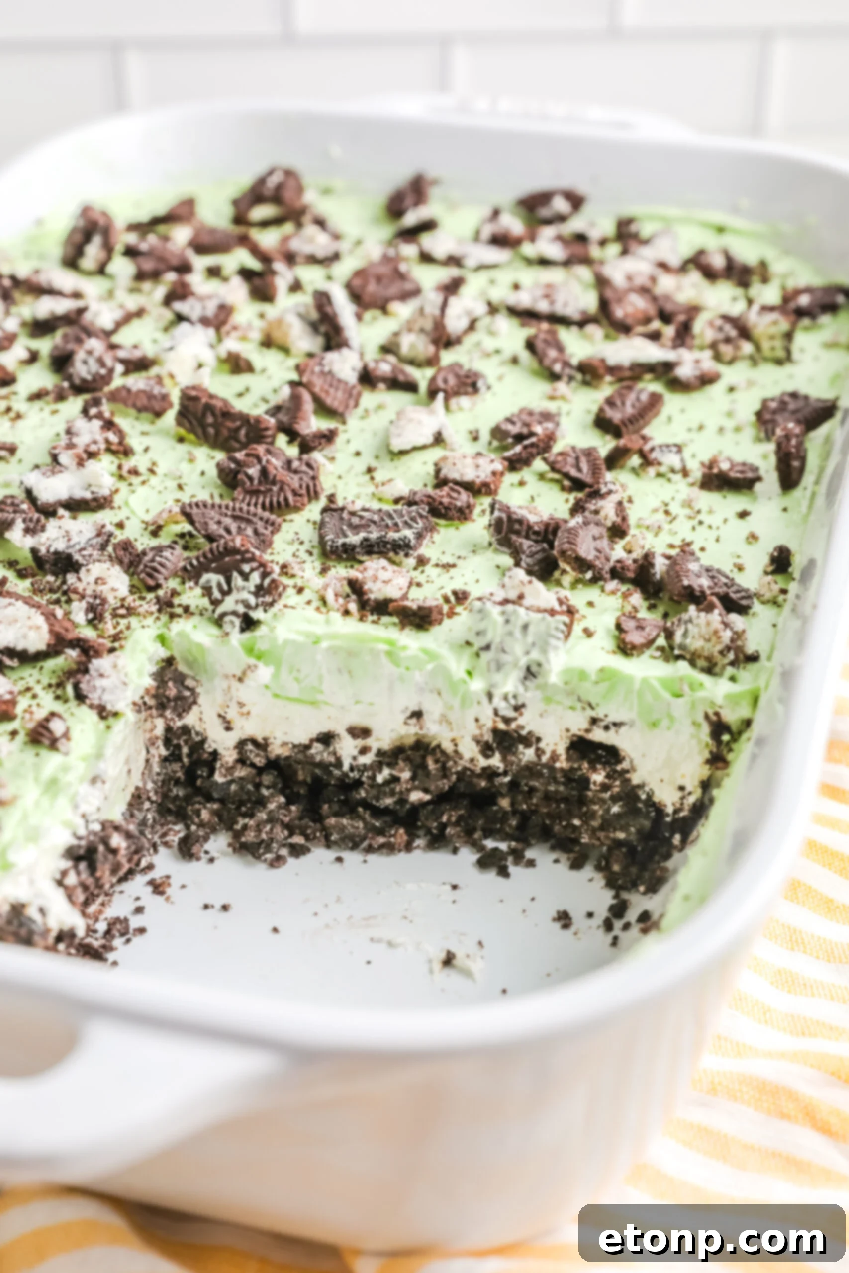 A slice of Shamrock Lasagna proudly displayed in a white baking dish, revealing its distinct, beautiful layers: the dark Oreo crust, the creamy white cheese layer, and the vibrant green mint pudding layer.