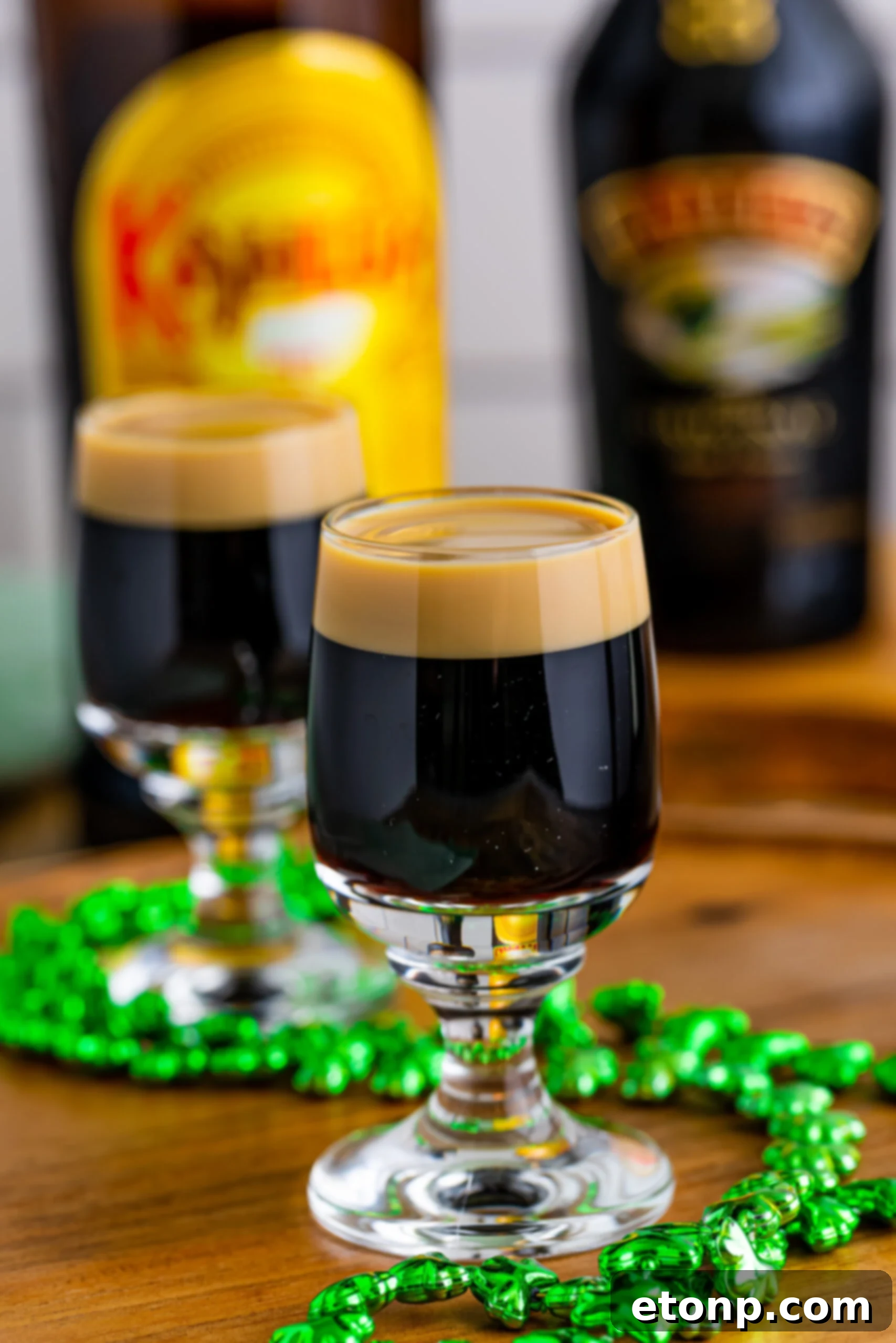 Two perfectly layered Baby Guinness Shots presented on a rustic wooden board, adorned with a festive green shamrock necklace, ready for a celebration.