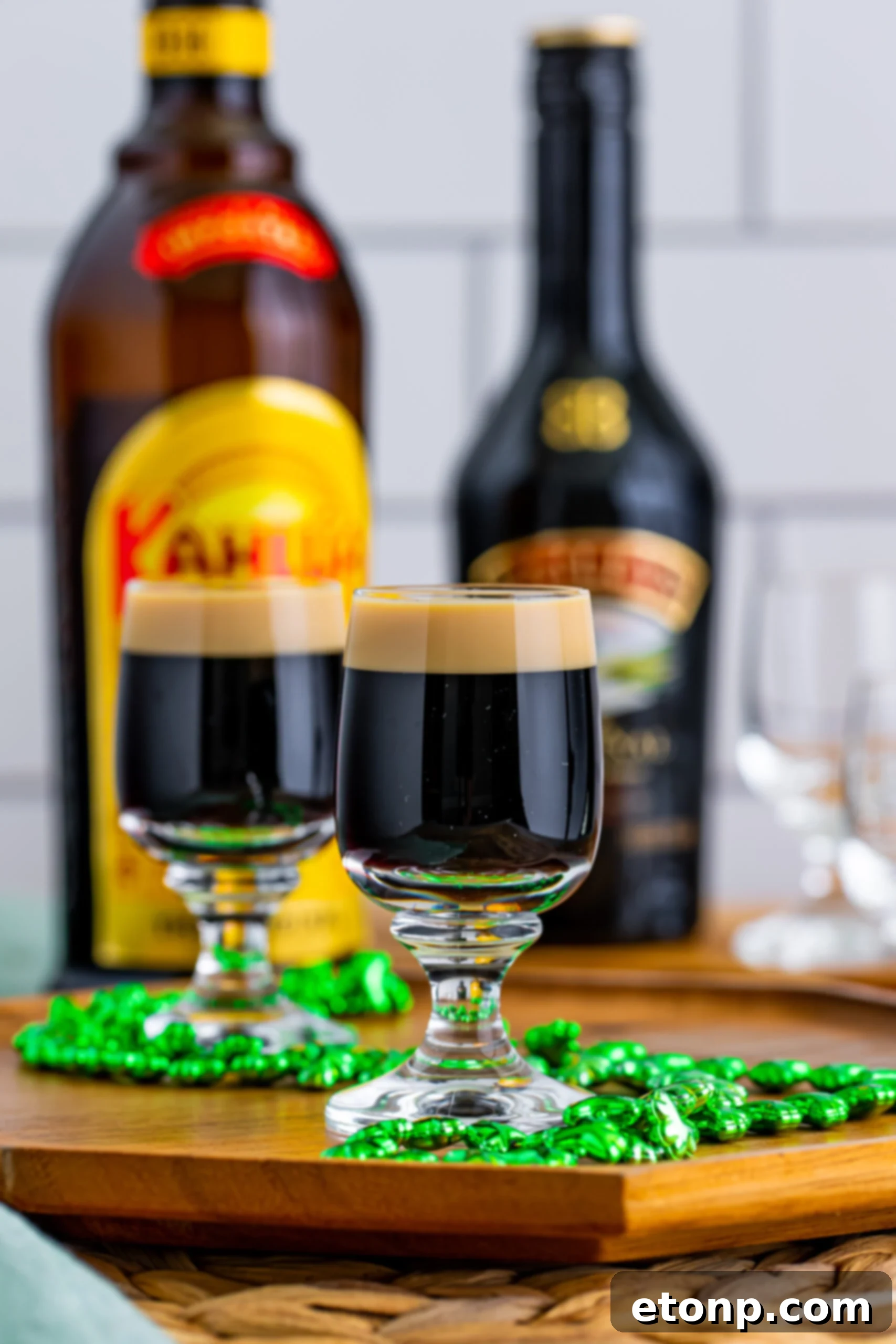 A close-up view of two Baby Guinness Shots, perfectly layered with dark Kahlua and a frothy head of Bailey's, sitting on a wooden serving board with a shamrock necklace.