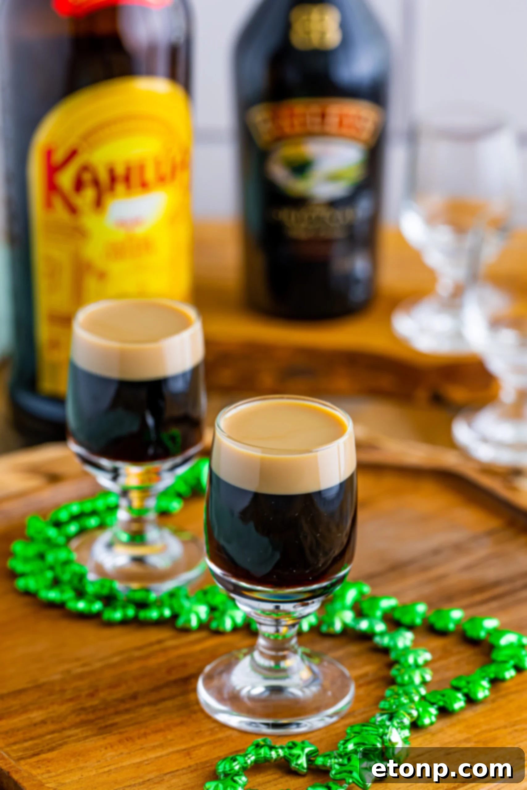 A pair of beautifully crafted Baby Guinness Shots, with their iconic dark and white layers, sitting prominently on a wooden surface beside a festive green shamrock necklace.