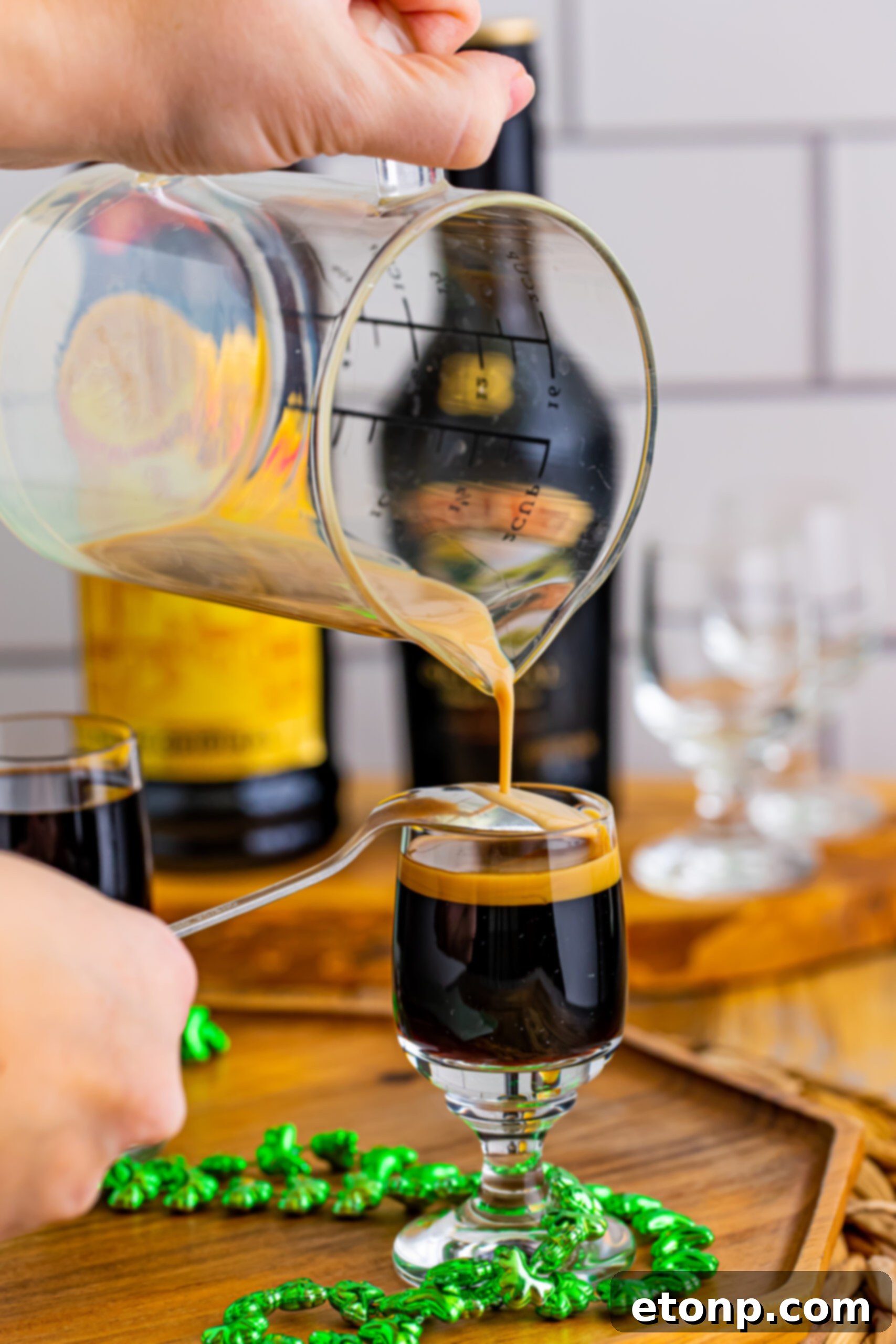A hand expertly pouring Bailey's Irish Cream over an inverted spoon into a shot glass already containing Kahlua, demonstrating the crucial layering technique for a Baby Guinness Shot.