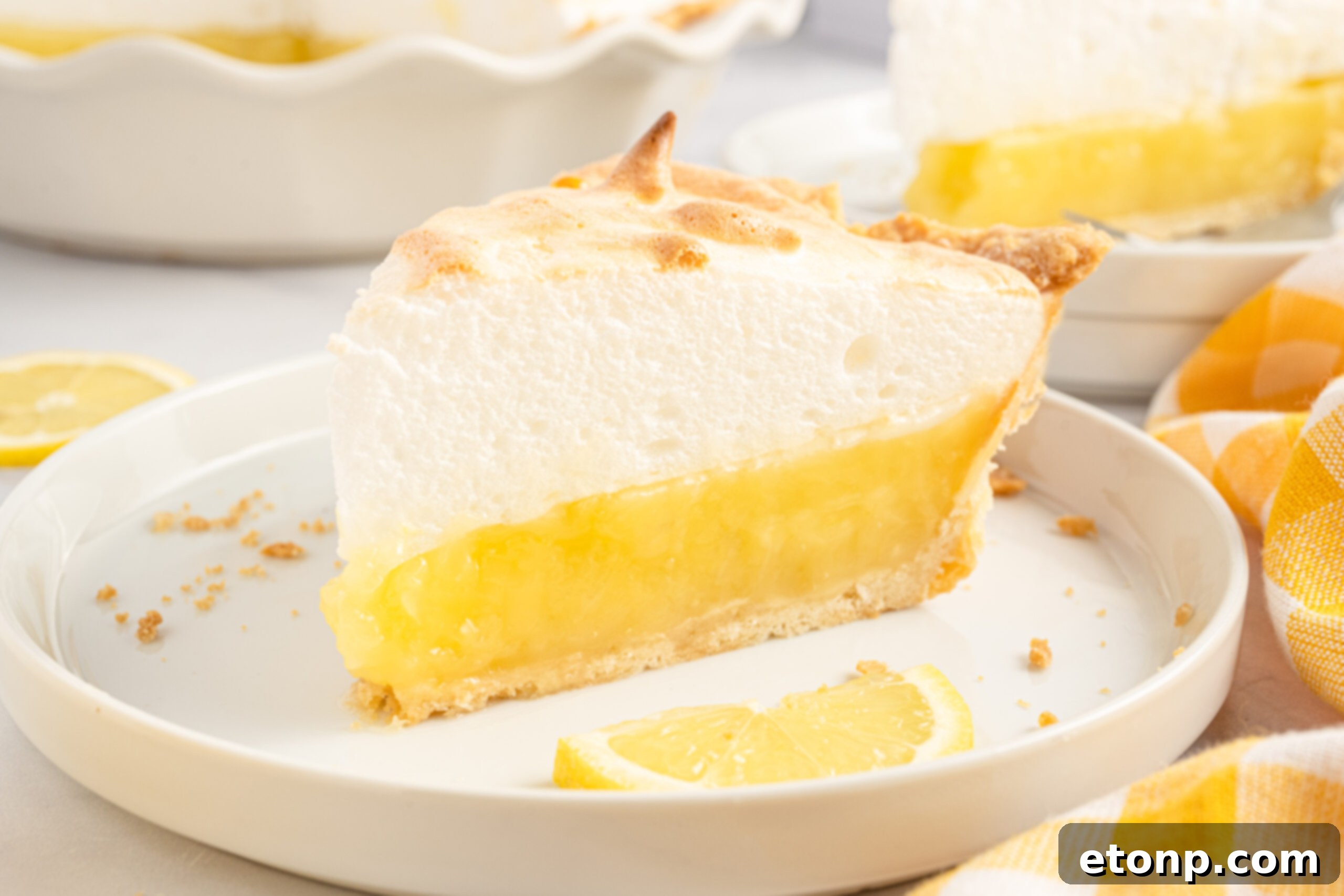 Side view of a slice of lemon meringue pie on a white plate, beautifully presented with fresh lemon slices.