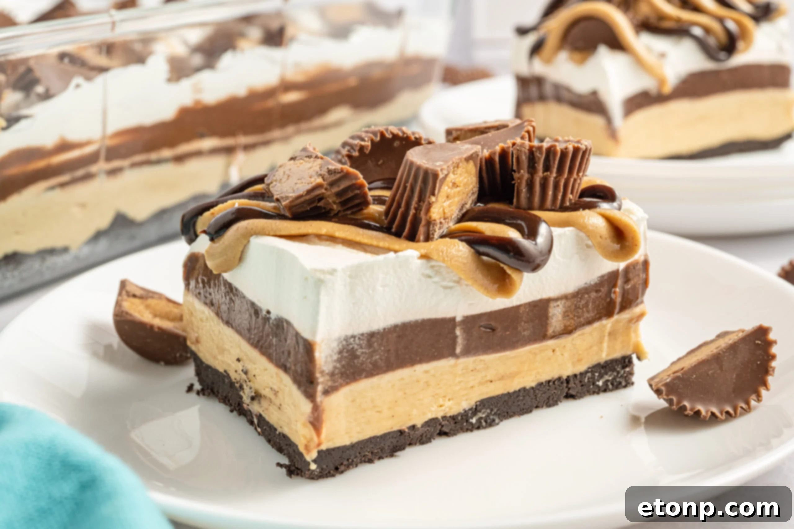 A piece of chocolate peanut butter lasagna on a white plate with Reese's cups in the background. 