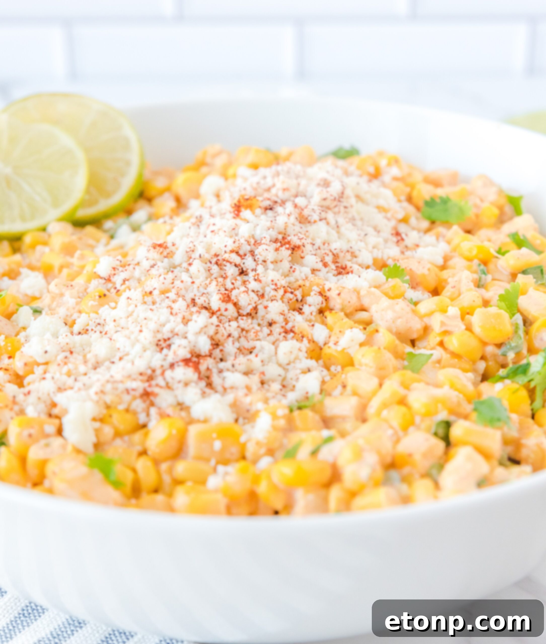 A close-up view of Mexican Street Corn Salad in a large white serving bowl, richly topped with crumbled feta cheese and bright lime slices.