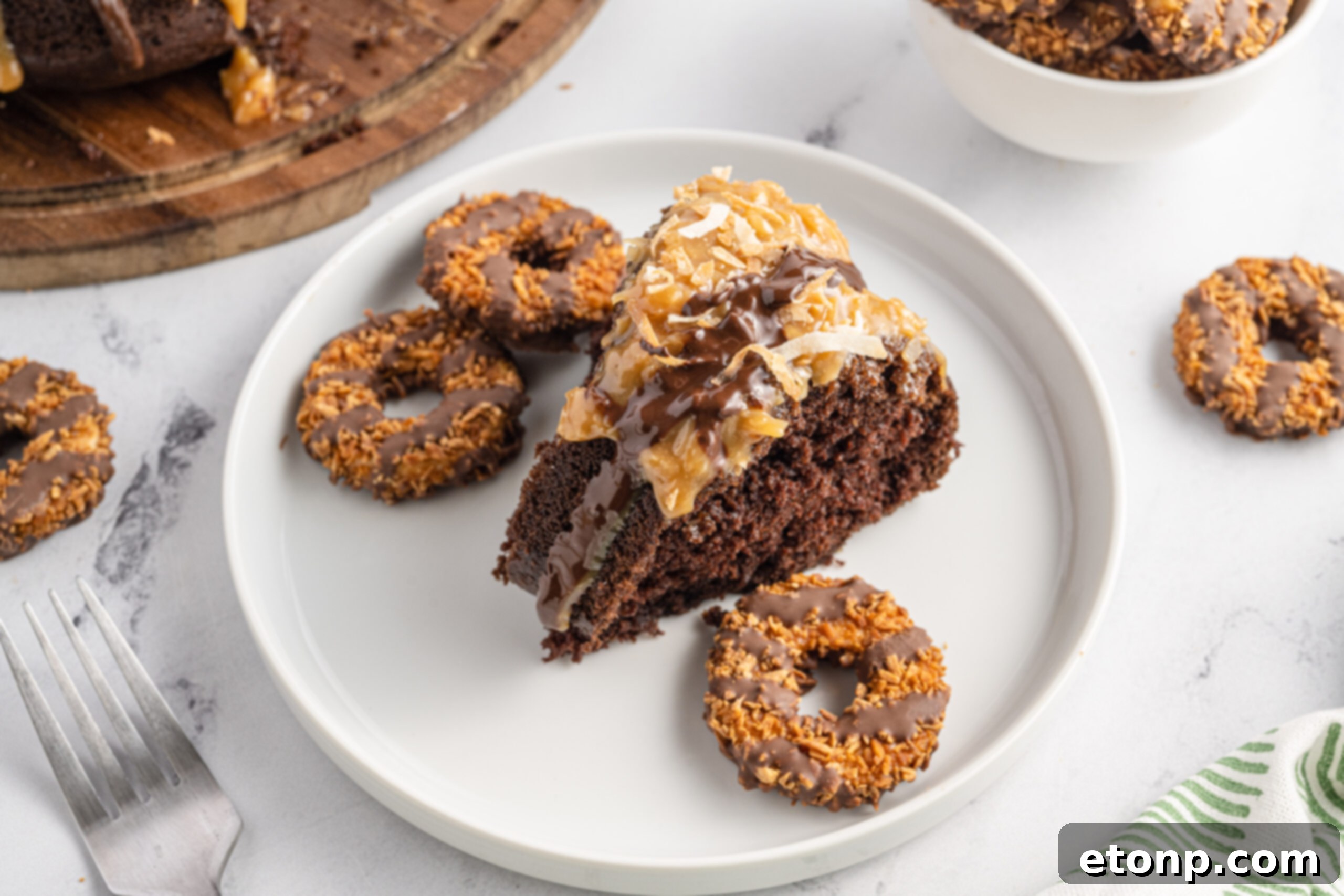A close-up view of a perfect slice of Samoa Cake served on a white plate, accompanied by a few classic Samoa Girl Scout cookies for a delightful comparison.