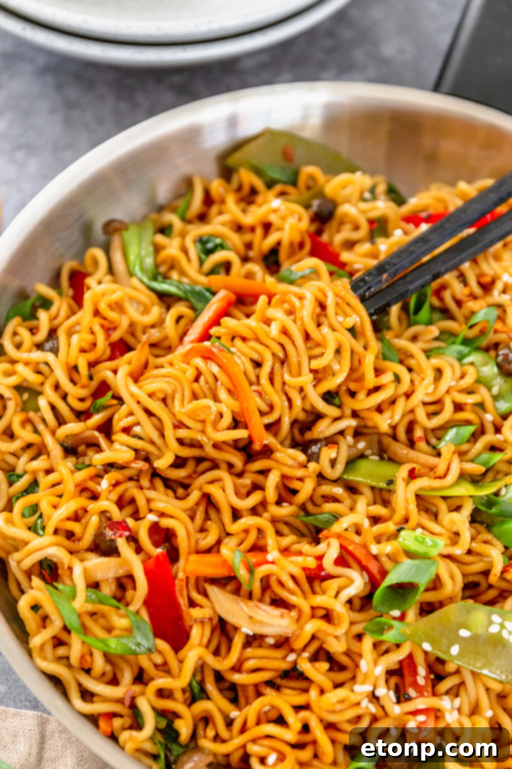 A pair of chopsticks lifting flavorful Ramen Stir Fry from a gleaming silver frying pan, showcasing tender noodles and colorful vegetables.