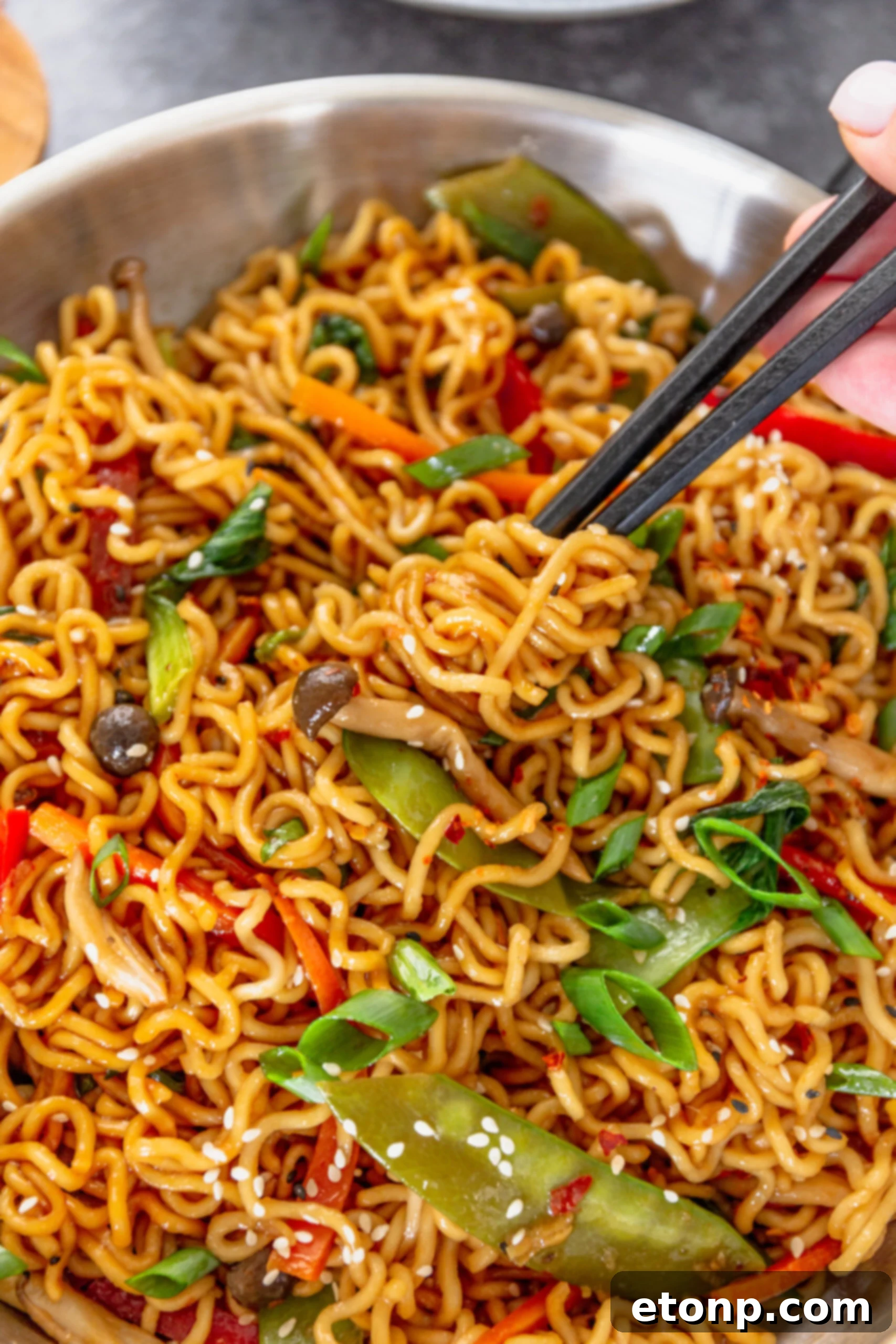 Chopsticks gracefully lifting a generous portion of Ramen Stir Fry from a shimmering silver frying pan, emphasizing its fresh ingredients.