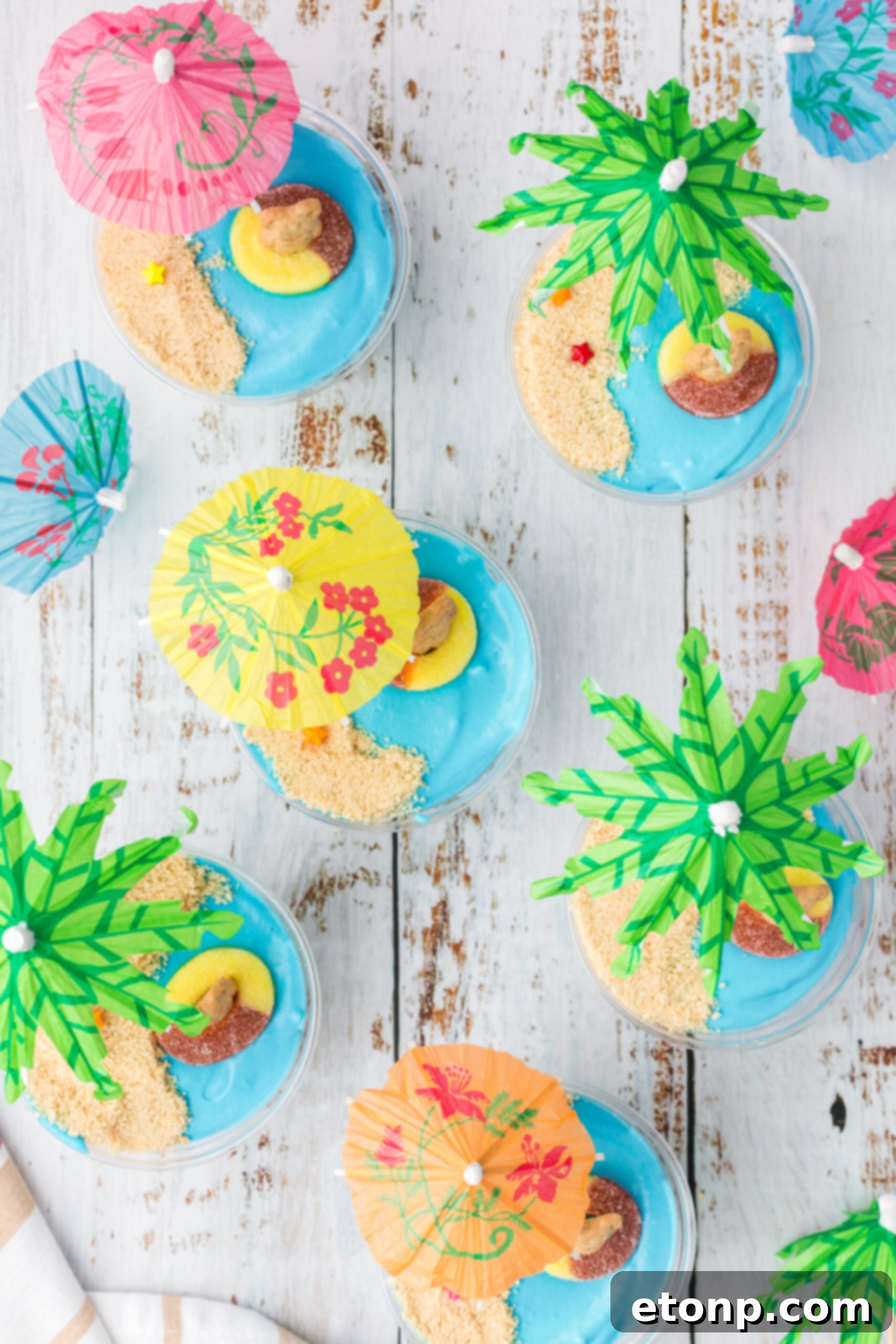 Overhead view of Beach Cheesecake Pudding Cups on a rustic wooden surface, with a toy shovel and seashells for decoration.