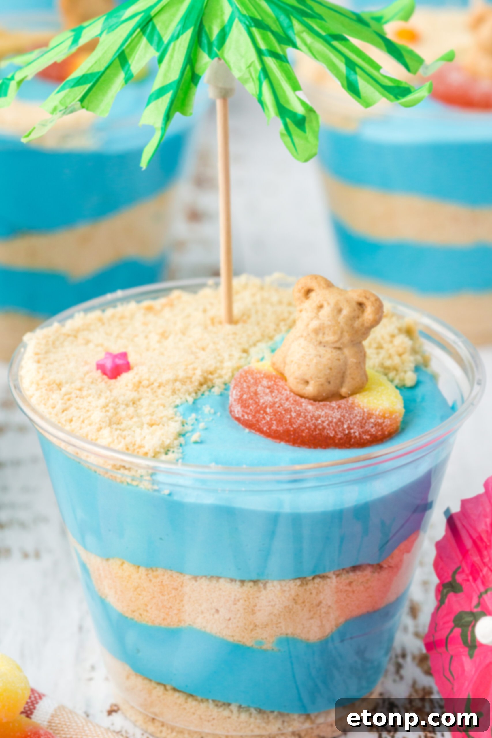 Close-up view of a single Beach Cheesecake Pudding Cup, highlighting the layers of 'sand' and blue 'ocean' pudding, adorned with a gummy ring and Teddy Graham.