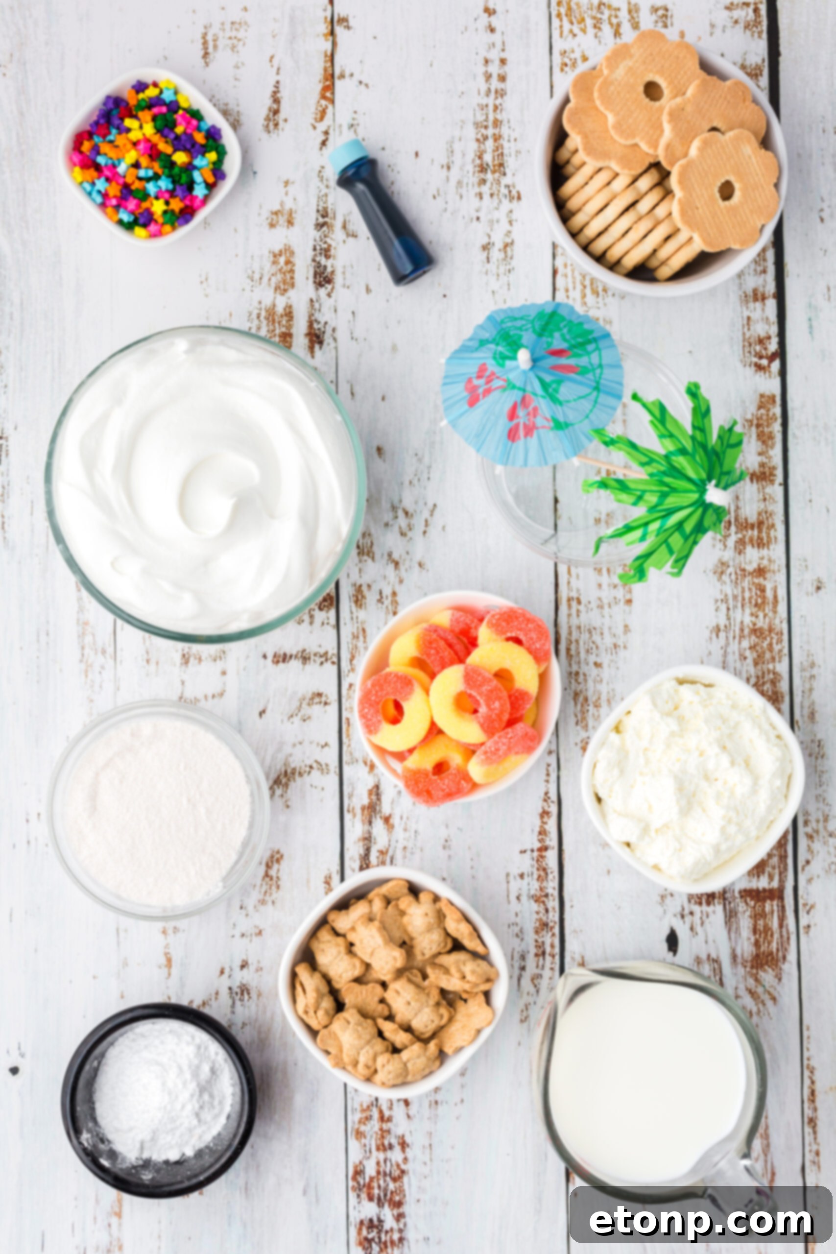 Overhead view of all ingredients for Beach Cheesecake Pudding Cups laid out on a white surface, including cookies, Cool Whip, cream cheese, pudding mix, milk, and various candies.