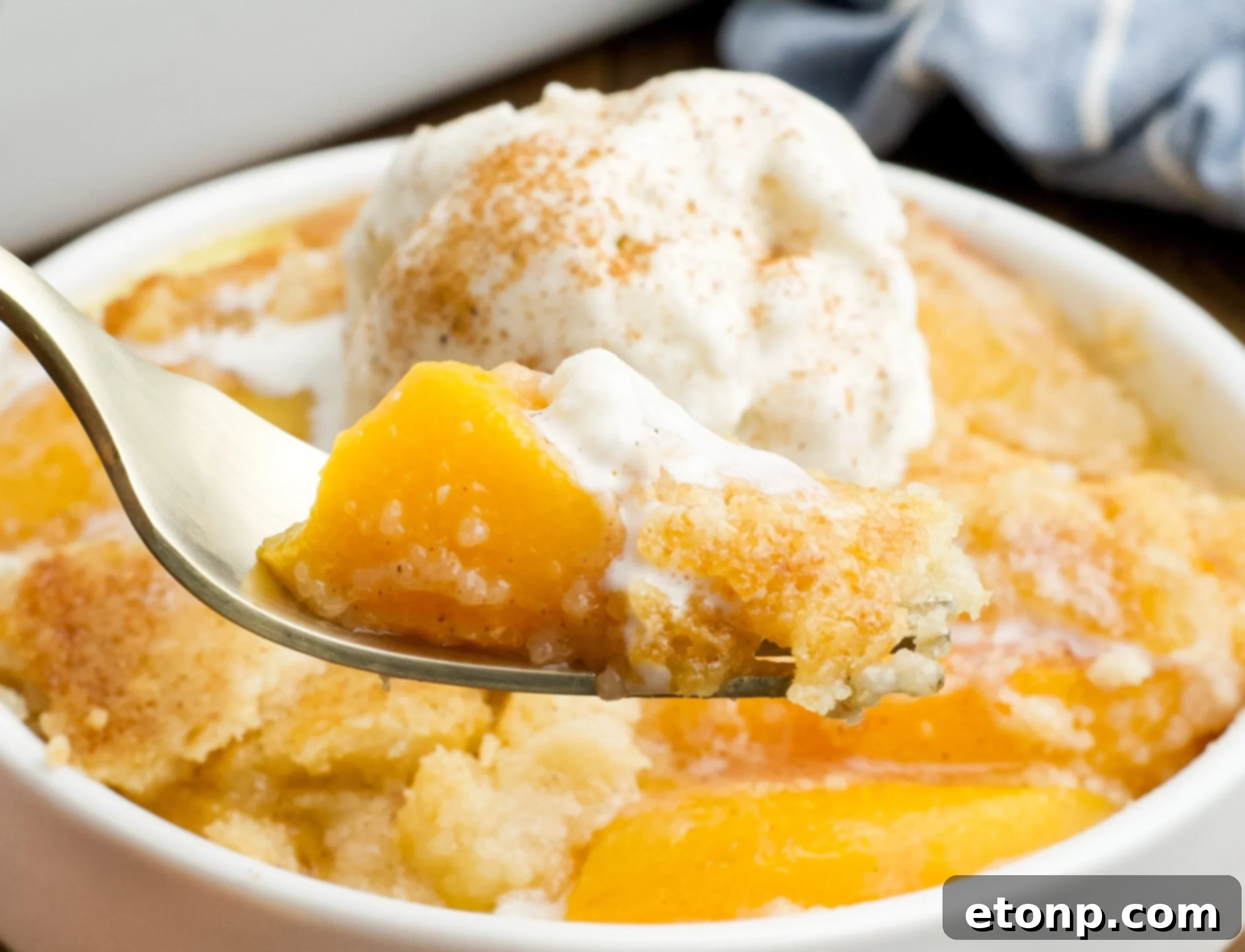 close up of of a serving of Peach Dump Cake in a white bowl with a gold fork removing a bite.