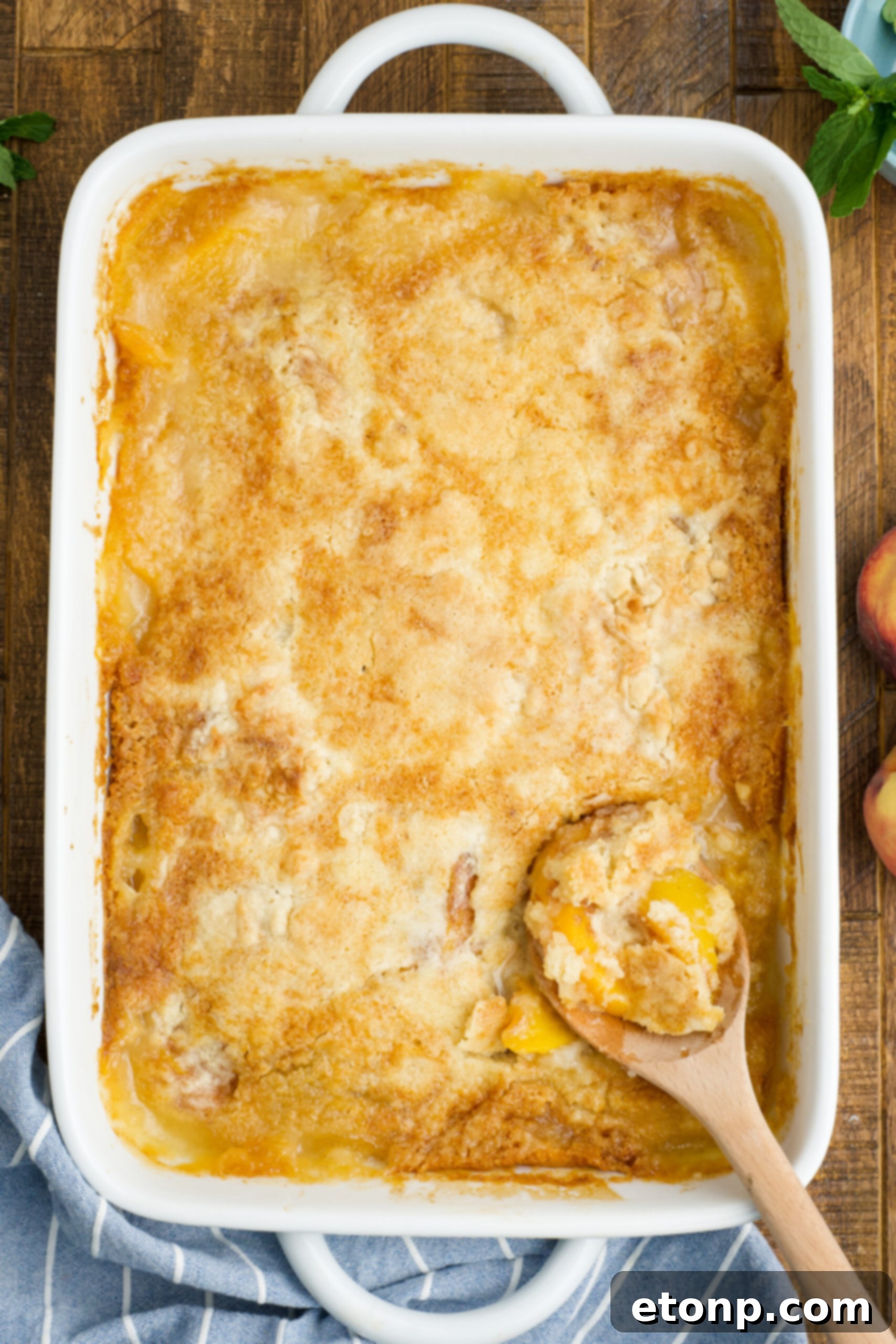Overhead view of Peach Dump Cake in a white baking dish with a wooden spoon removing a serving.