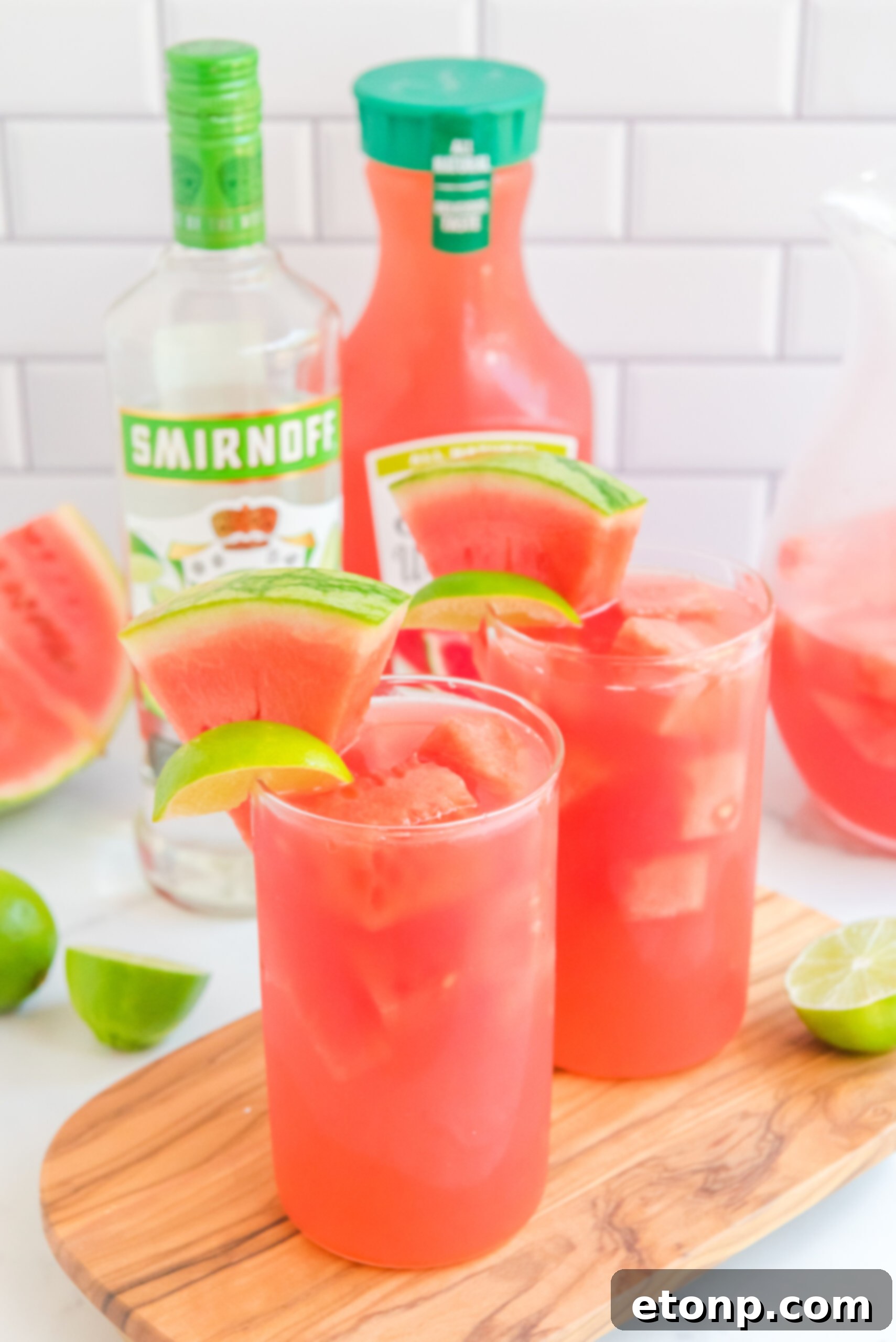 Front view of Watermelon Cocktail in two clear glasses garnished with lime and watermelon wedges on a wooden board.
