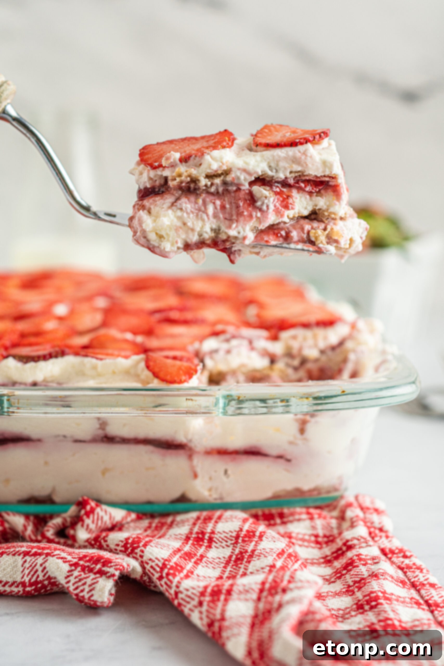 A glorious Strawberry Cheesecake Icebox Cake in a baking dish, with a slice being lifted by a silver spatula, revealing its beautiful layers.