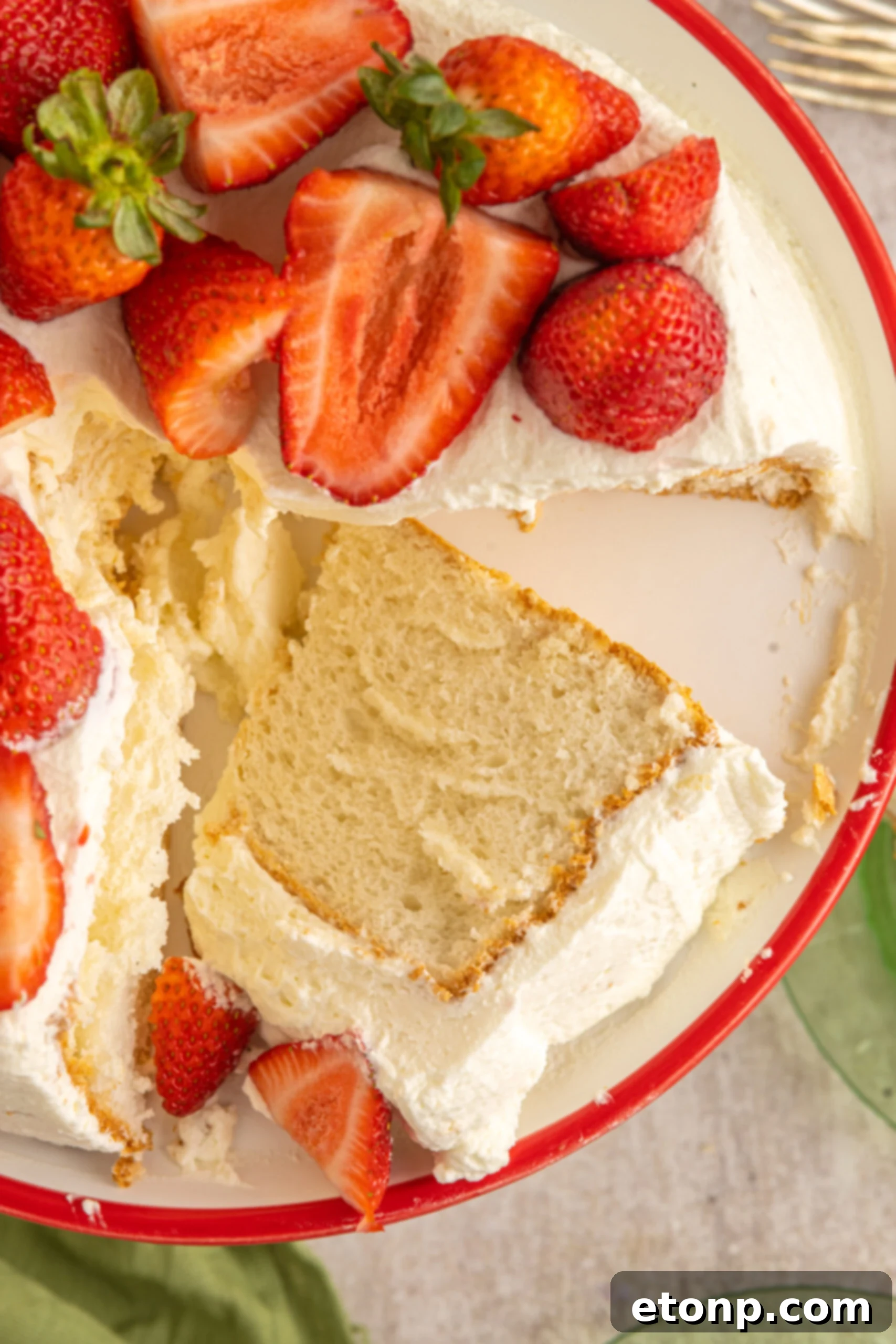 An overhead view of a beautifully cut piece of Strawberry Angel Food Cake, highlighting its airy texture and fresh strawberry topping on a white and red cake stand.