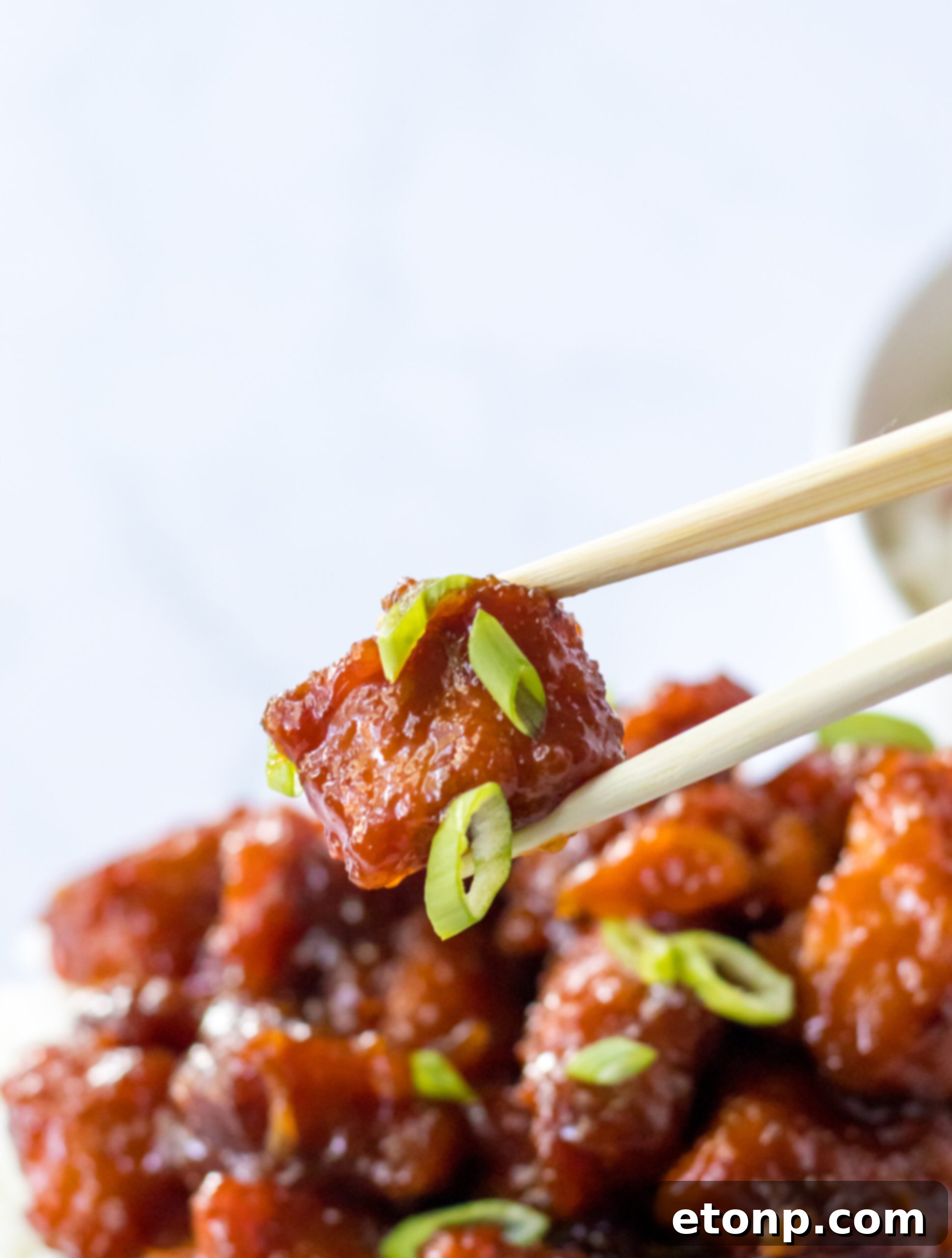 Close-up view of chopsticks holding a juicy piece of Slow Cooker Firecracker Chicken with a perfect glaze, against a blurred background.