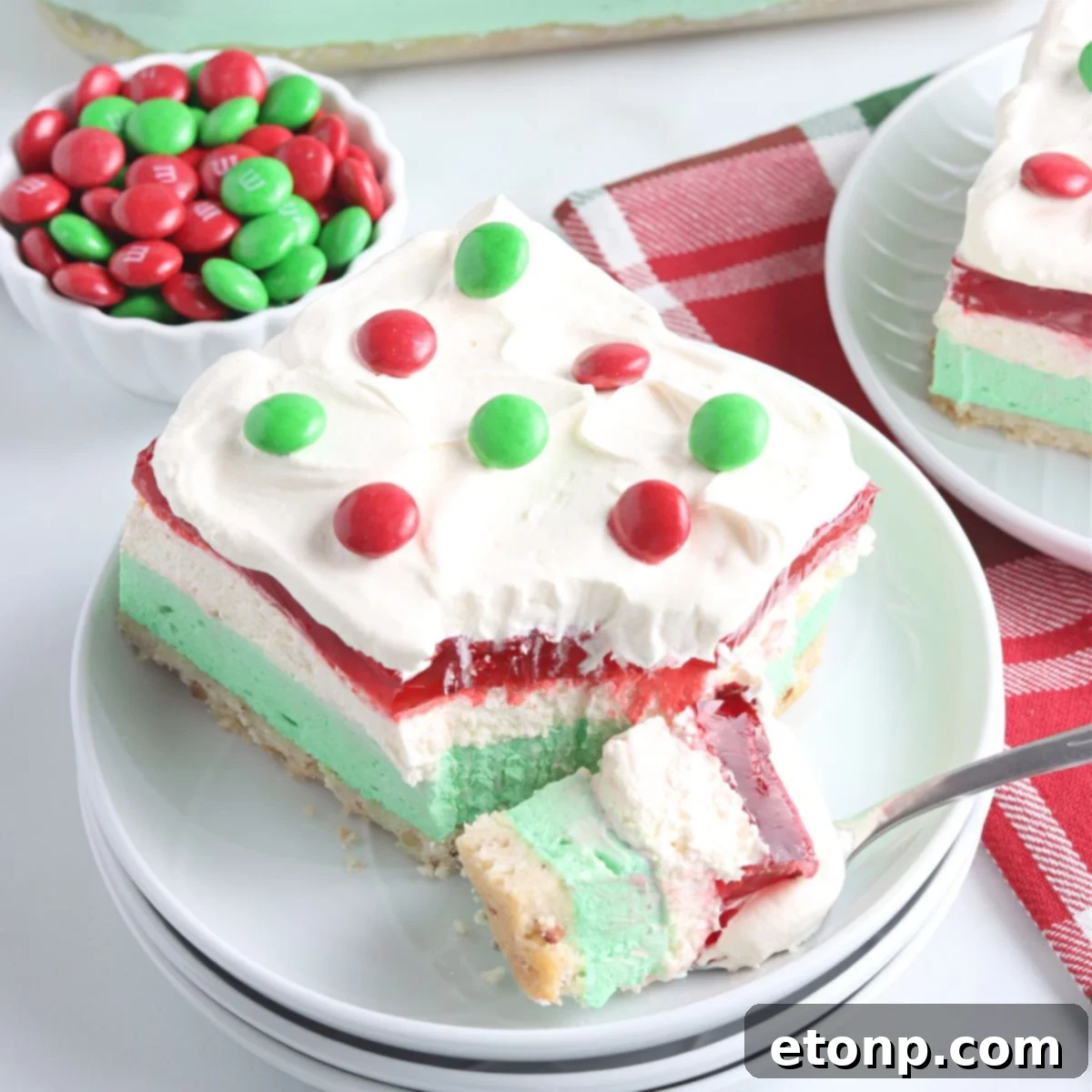 A perfectly sliced Christmas Lasagna dessert presented on a white plate, with a fork holding a bite-sized portion to showcase the vibrant layers of red, white, and green.