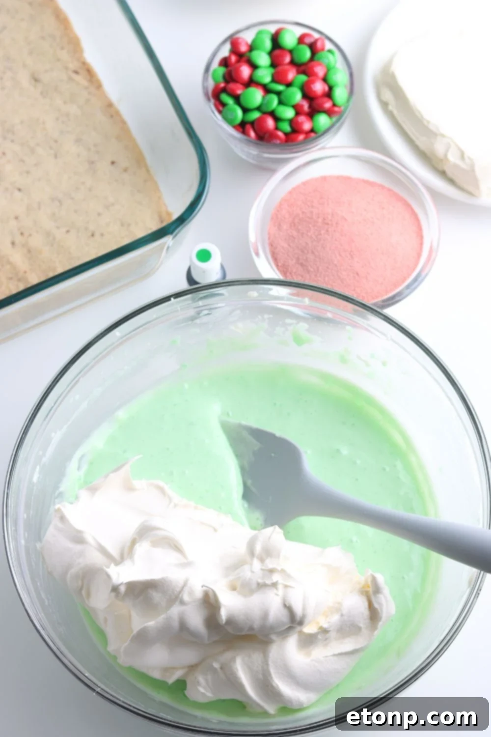 A hand gently folds whipped cream into a bright green jello mixture in a glass bowl, creating a light and airy consistency for a dessert layer.