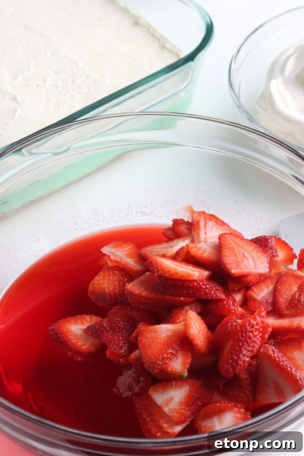 A bowl filled with sliced fresh strawberries gently mixed into a partially set strawberry jello, forming the final, fruity layer of the layered Christmas dessert.