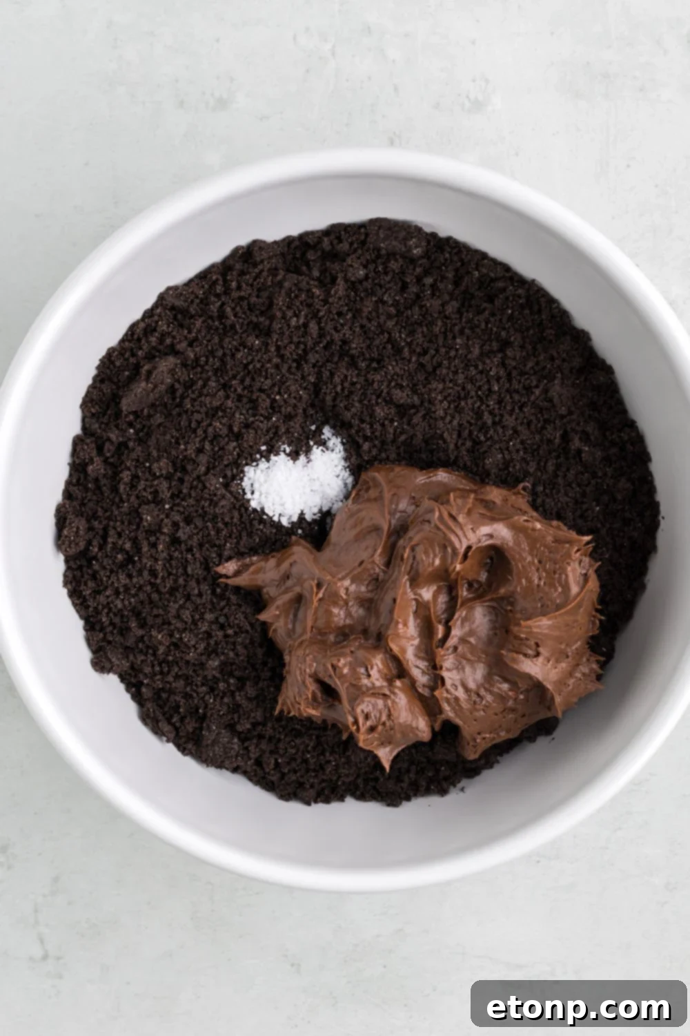 A bowl of crushed Oreos generously topped with rich chocolate frosting and a sprinkle of salt, awaiting thorough mixing.