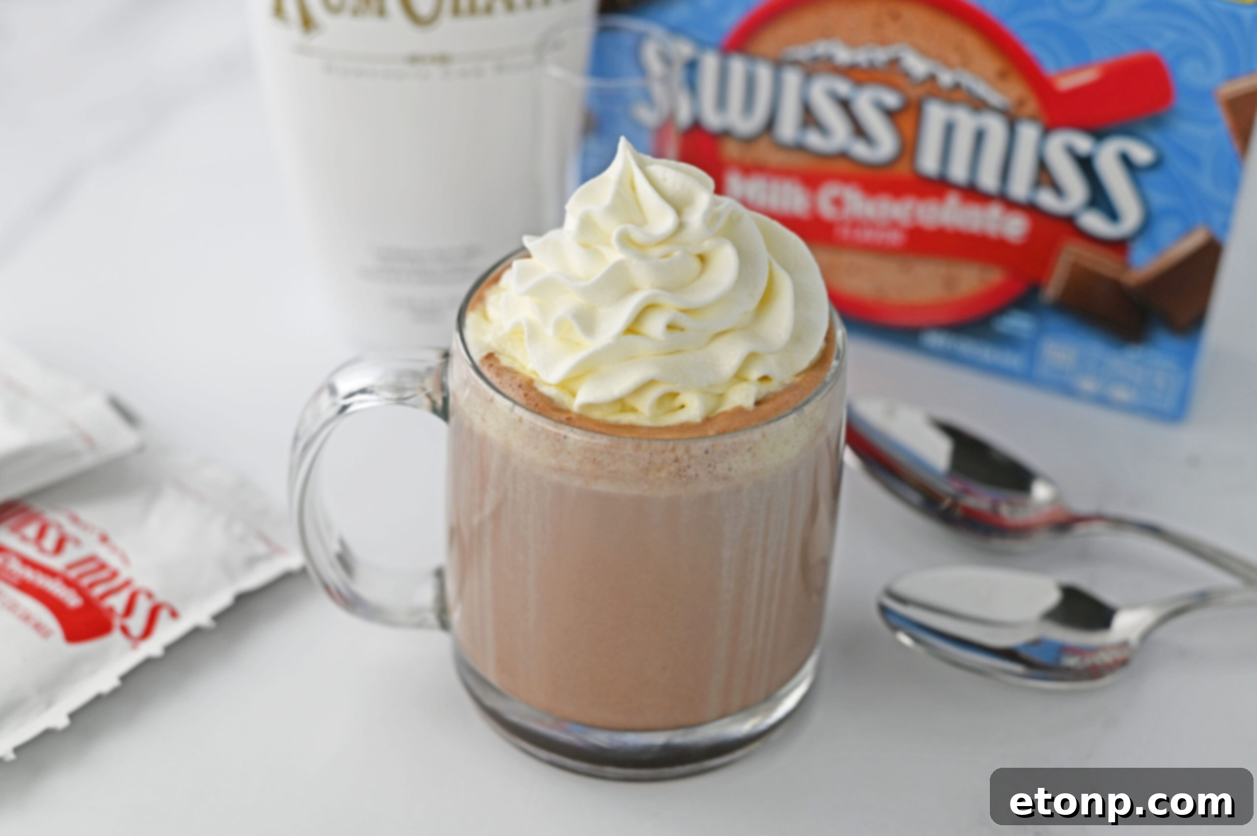 rumchata in a clear glass mug topped with whipped cream with a box of swiss miss and rumchata in the background