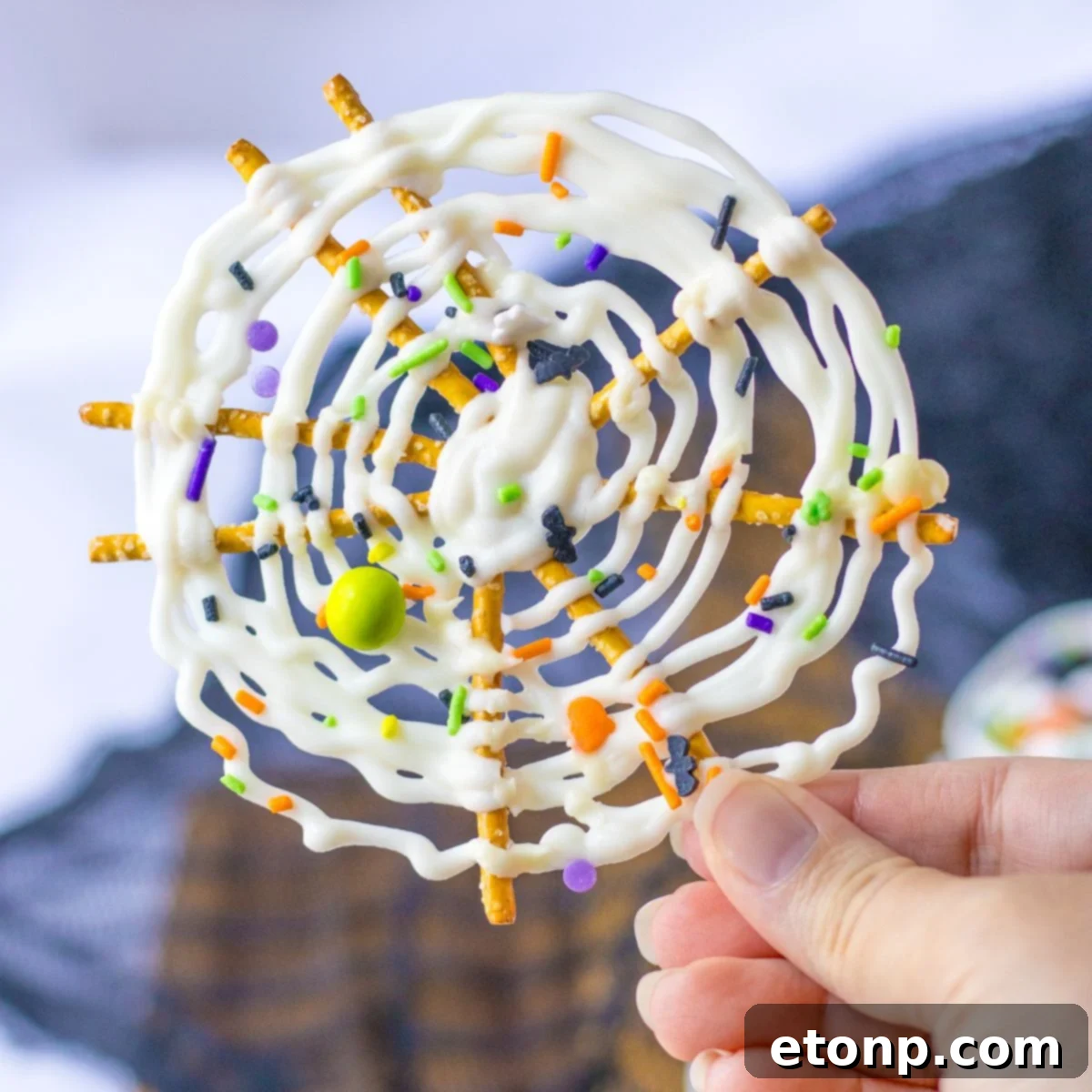 A hand holds a beautifully decorated Spider Web Pretzel with white chocolate and Halloween sprinkles