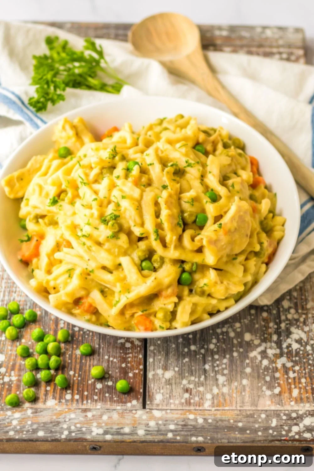 chicken noodles in a bowl with peas on table and wooden spoon behind it