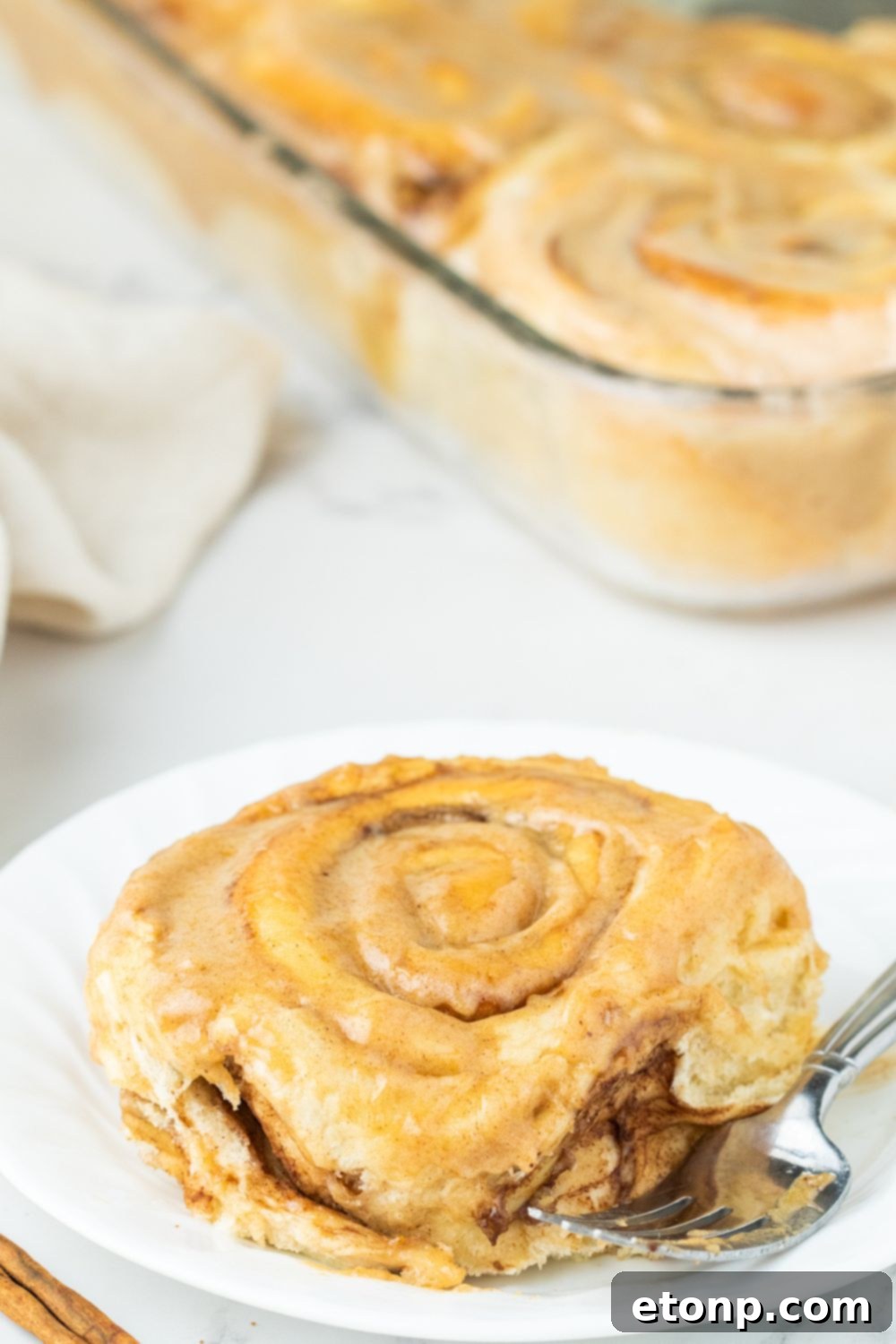 A frosted pumpkin cinnamon roll on a plate with a casserole dish of more rolls in the background