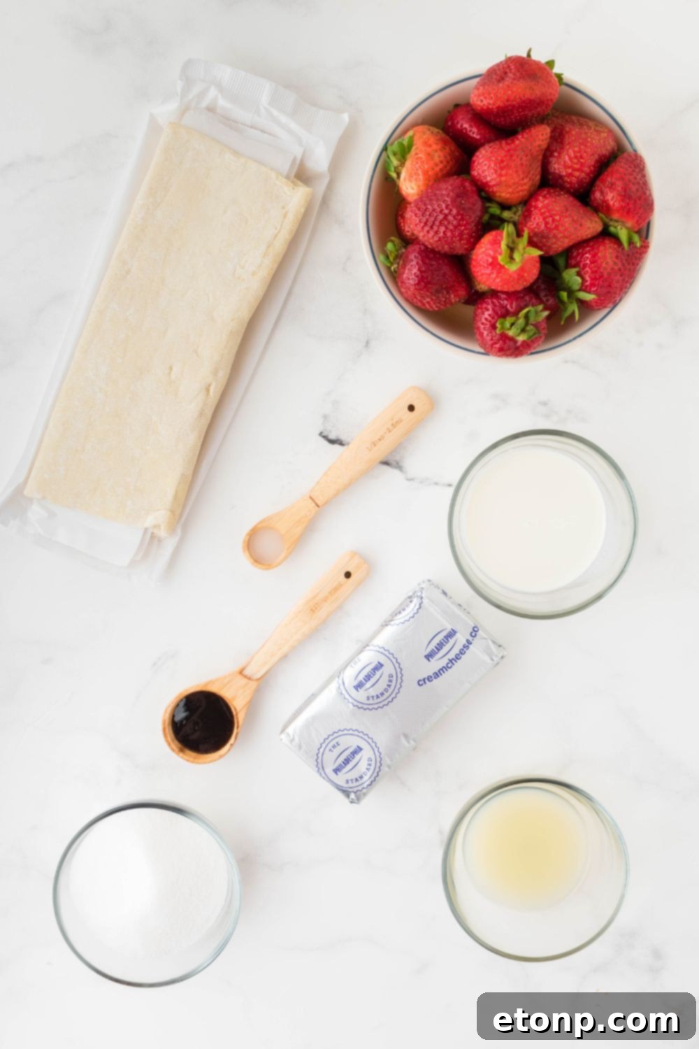 Arrangement of key puff pastry ingredients in elegant glass bowls on a table, including fresh strawberries, cream cheese, and various extracts.